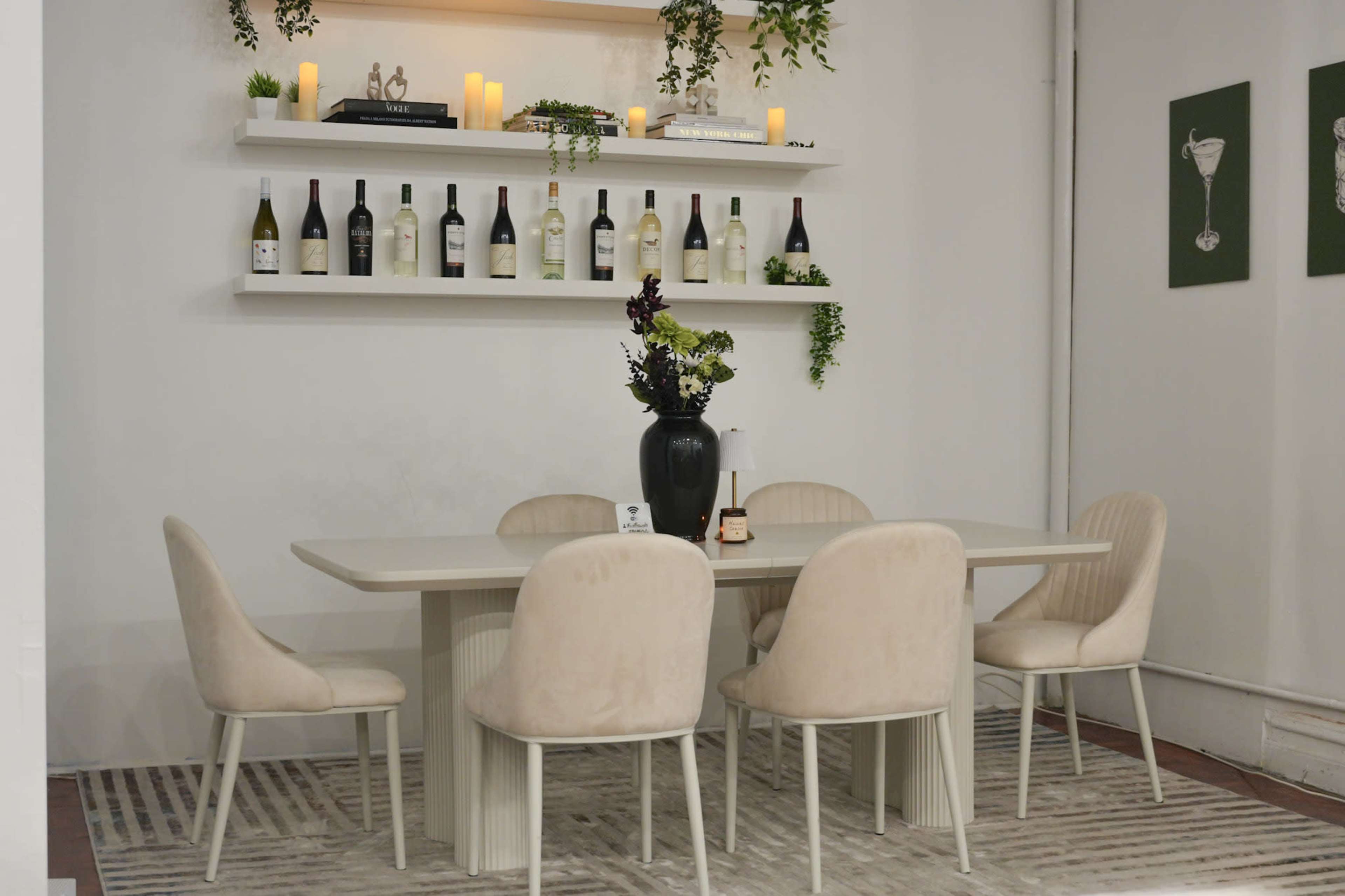 A dining area features a light-colored table surrounded by four chairs, with a shelf displaying wine bottles and green plants above.