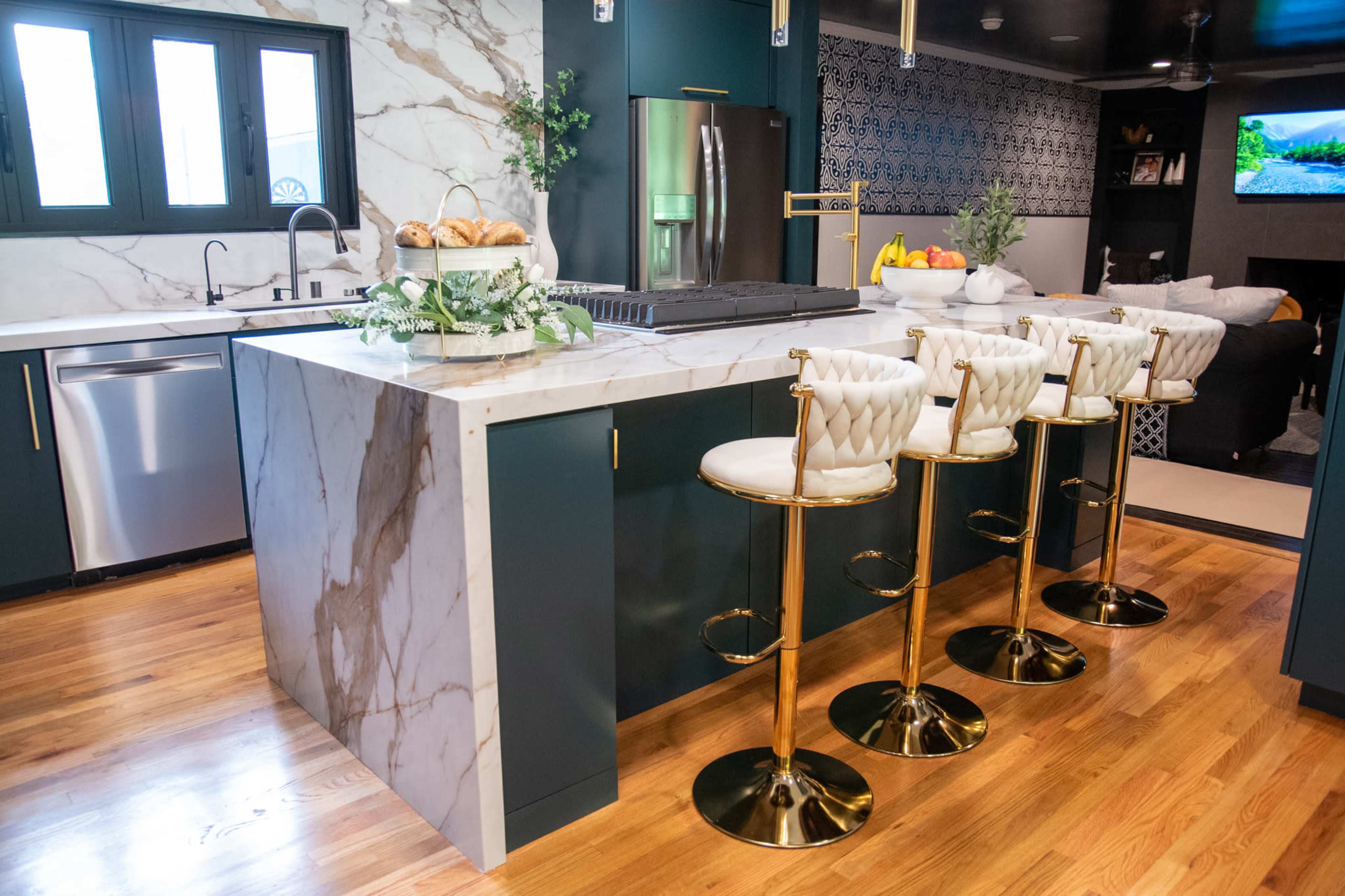 The image shows a modern kitchen with a marble island, gold bar stools, and various kitchen appliances, including a refrigerator and a stove.