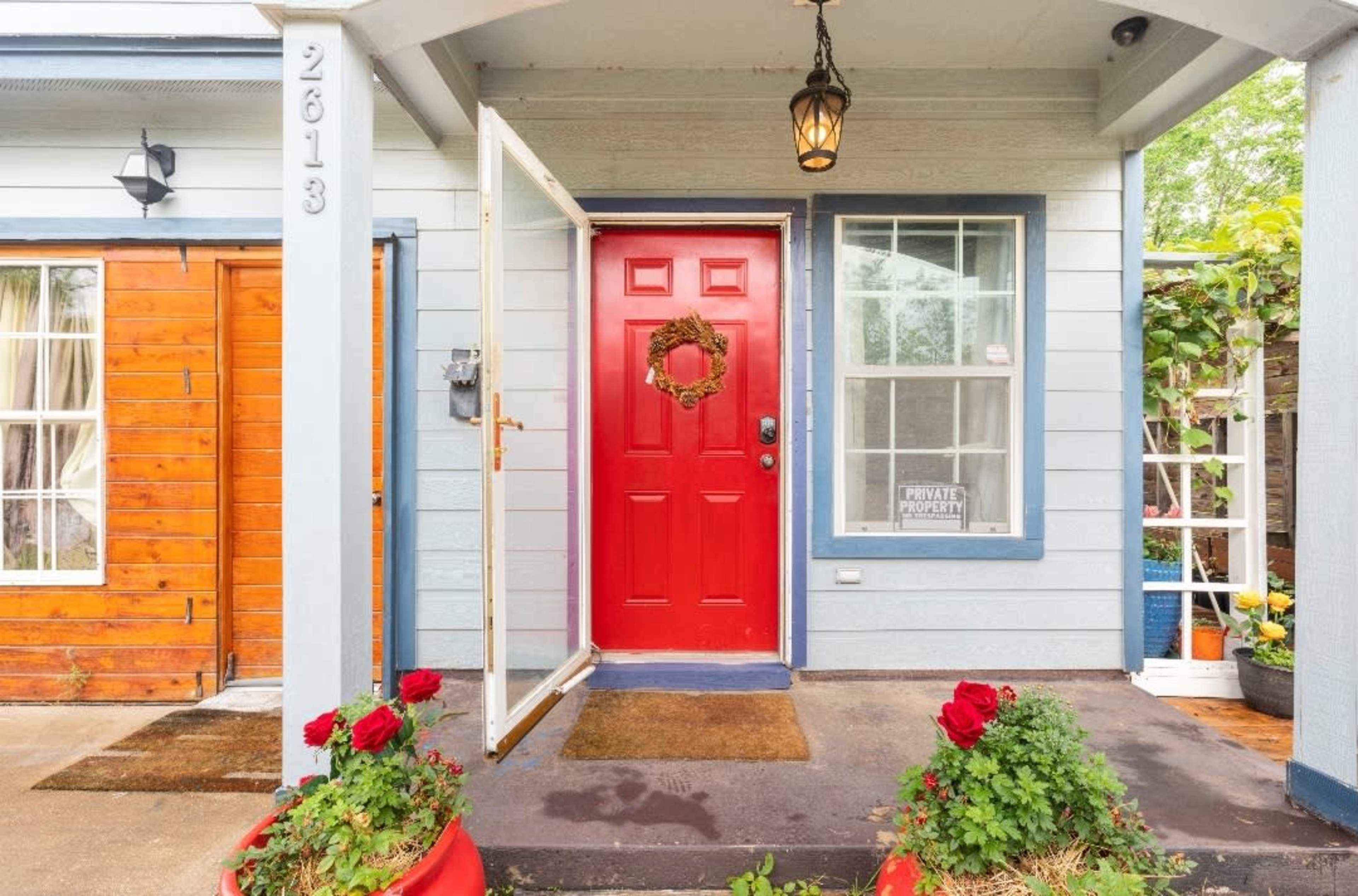 A red front door with a wreath is flanked by potted roses and leads to a house with blue siding.