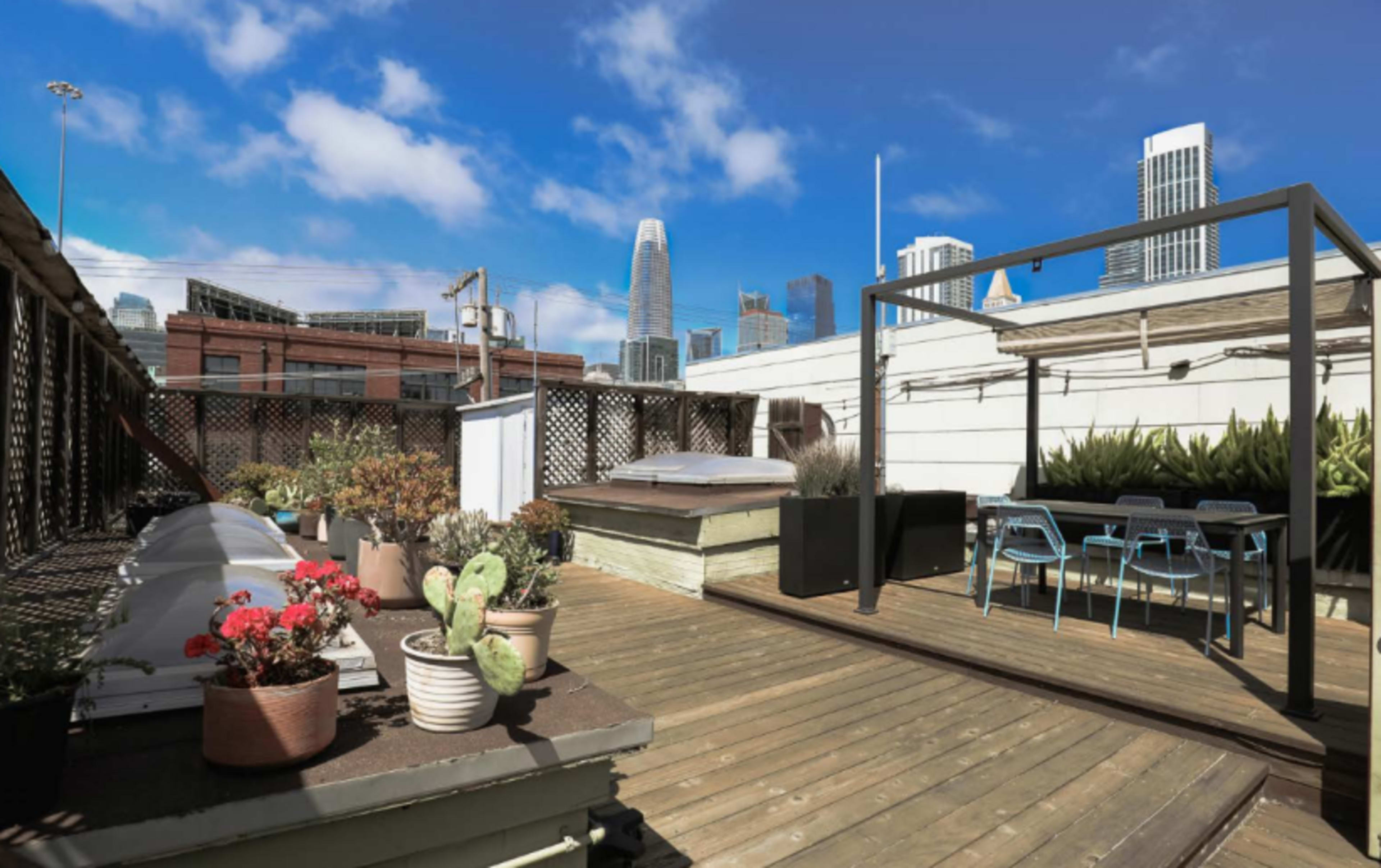 The image shows a rooftop terrace with wooden flooring, potted plants, a dining area with a table and chairs, and a view of skyscrapers in the background.