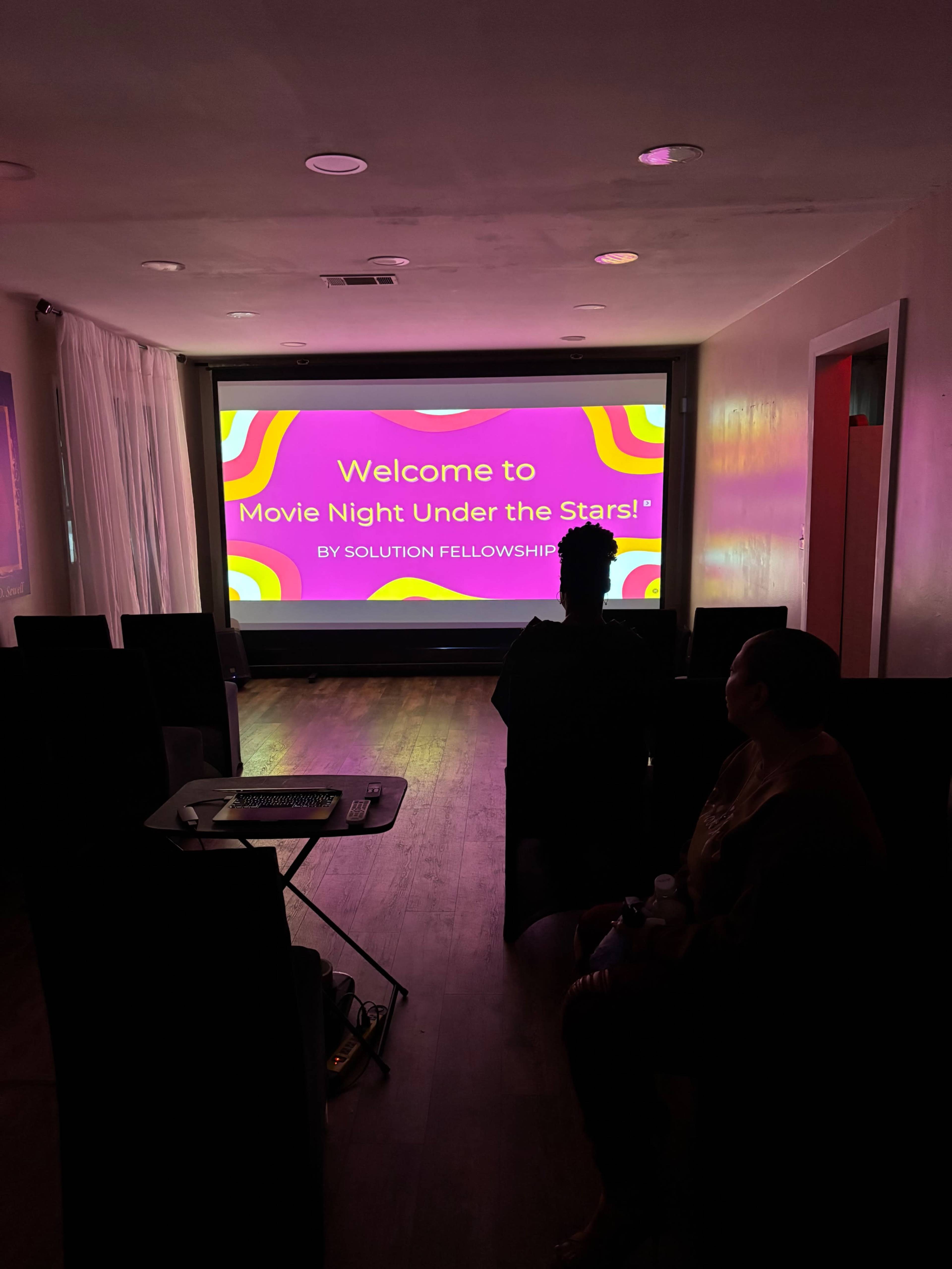 A cozy indoor setting features a large screen displaying "Welcome to Movie Night Under the Stars!" while two people sit in the foreground.