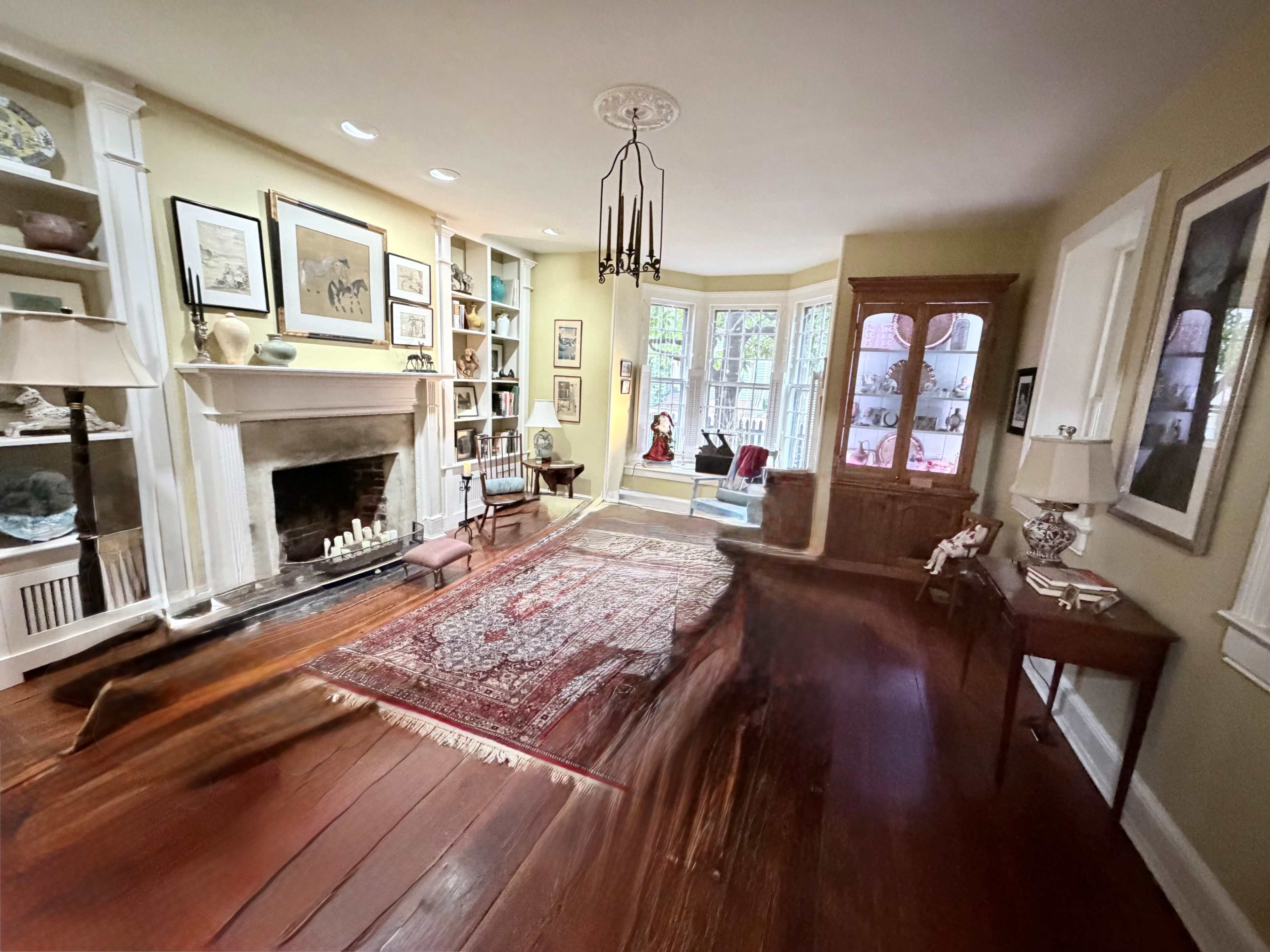 The image shows a living room with hardwood floors, a fireplace, built-in shelves, and large windows allowing natural light to enter.