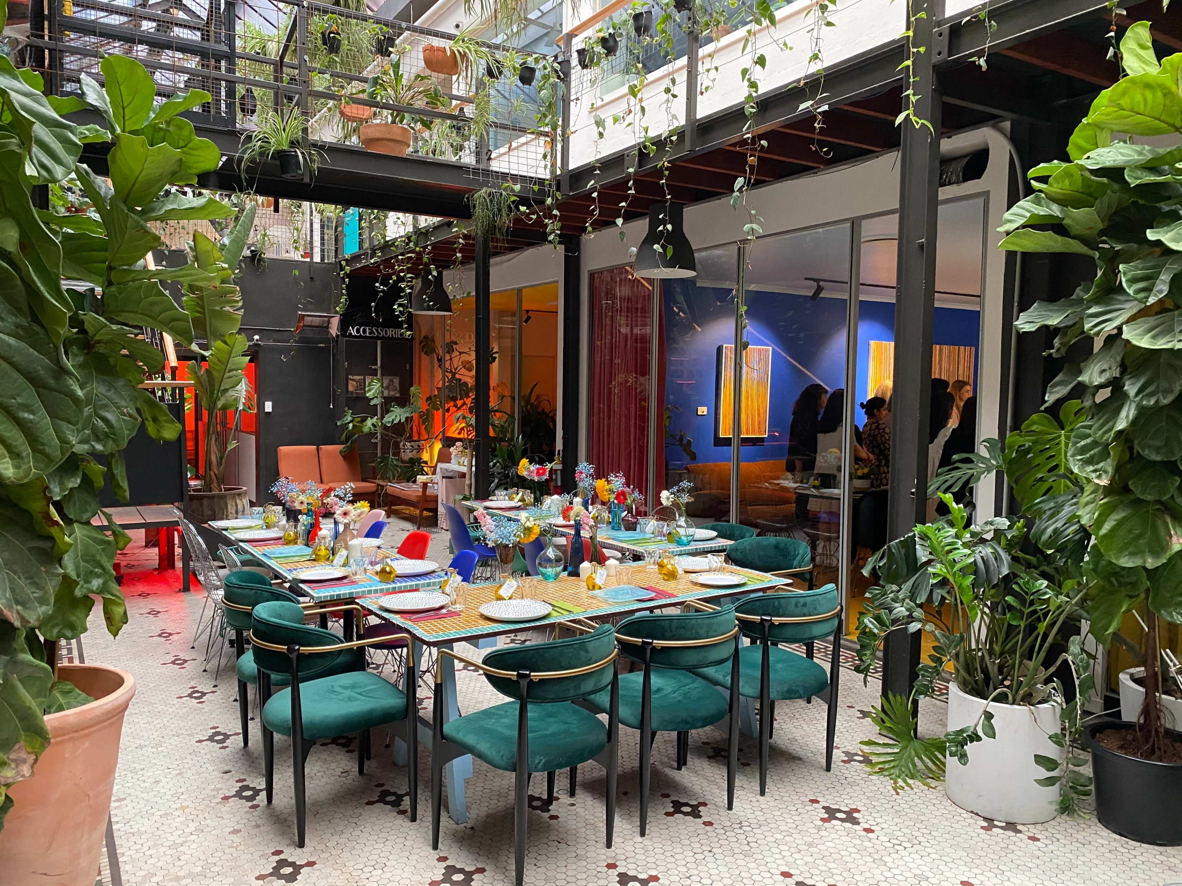 A long dining table is set with colorful tableware in a plant-filled indoor space with large windows and vibrant wall art.