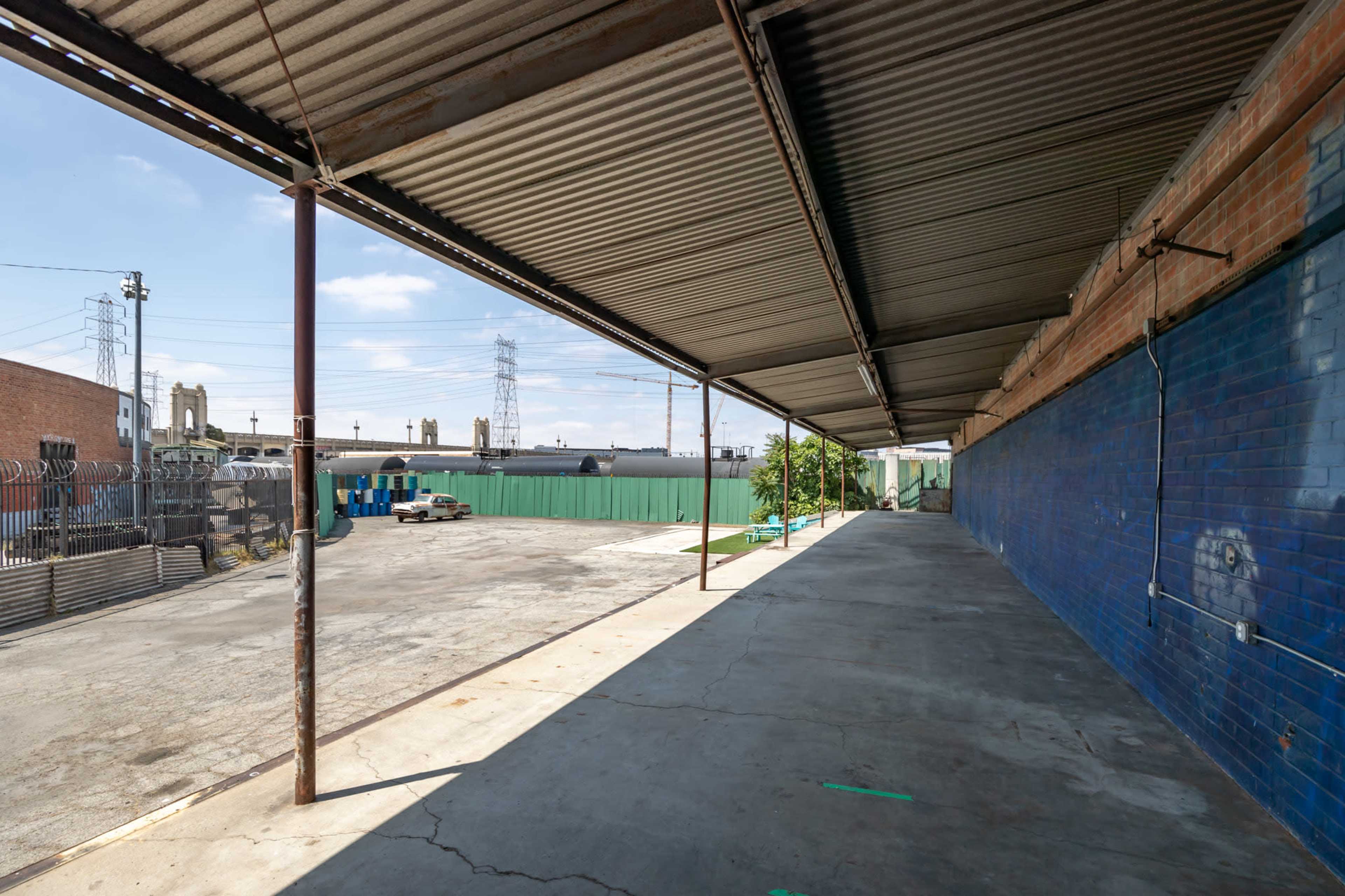 The image shows a covered walkway beside a concrete area with industrial buildings and green storage containers in the background.