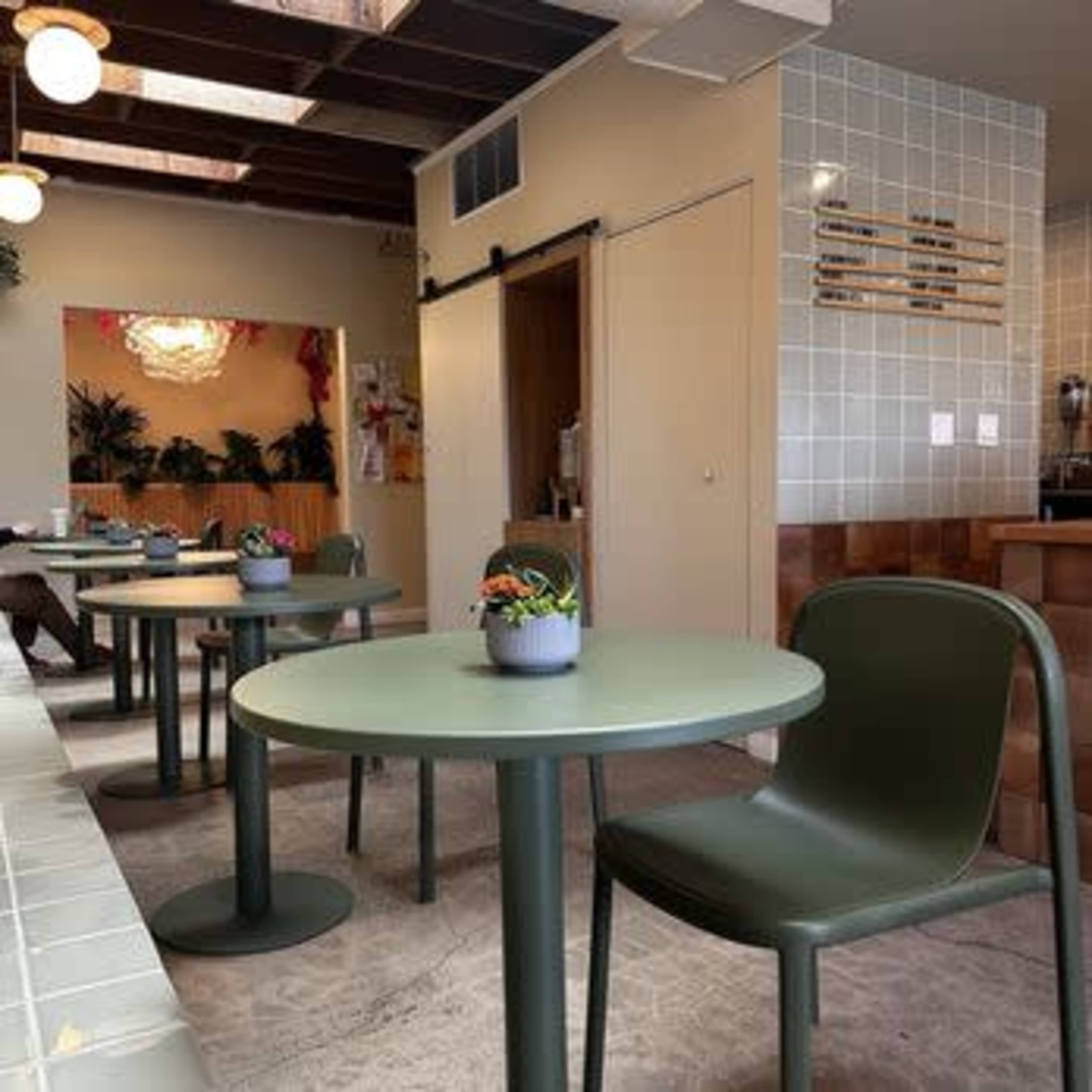 The image shows a modern cafe interior with green tables and chairs, potted plants on the tables, and tiled walls.