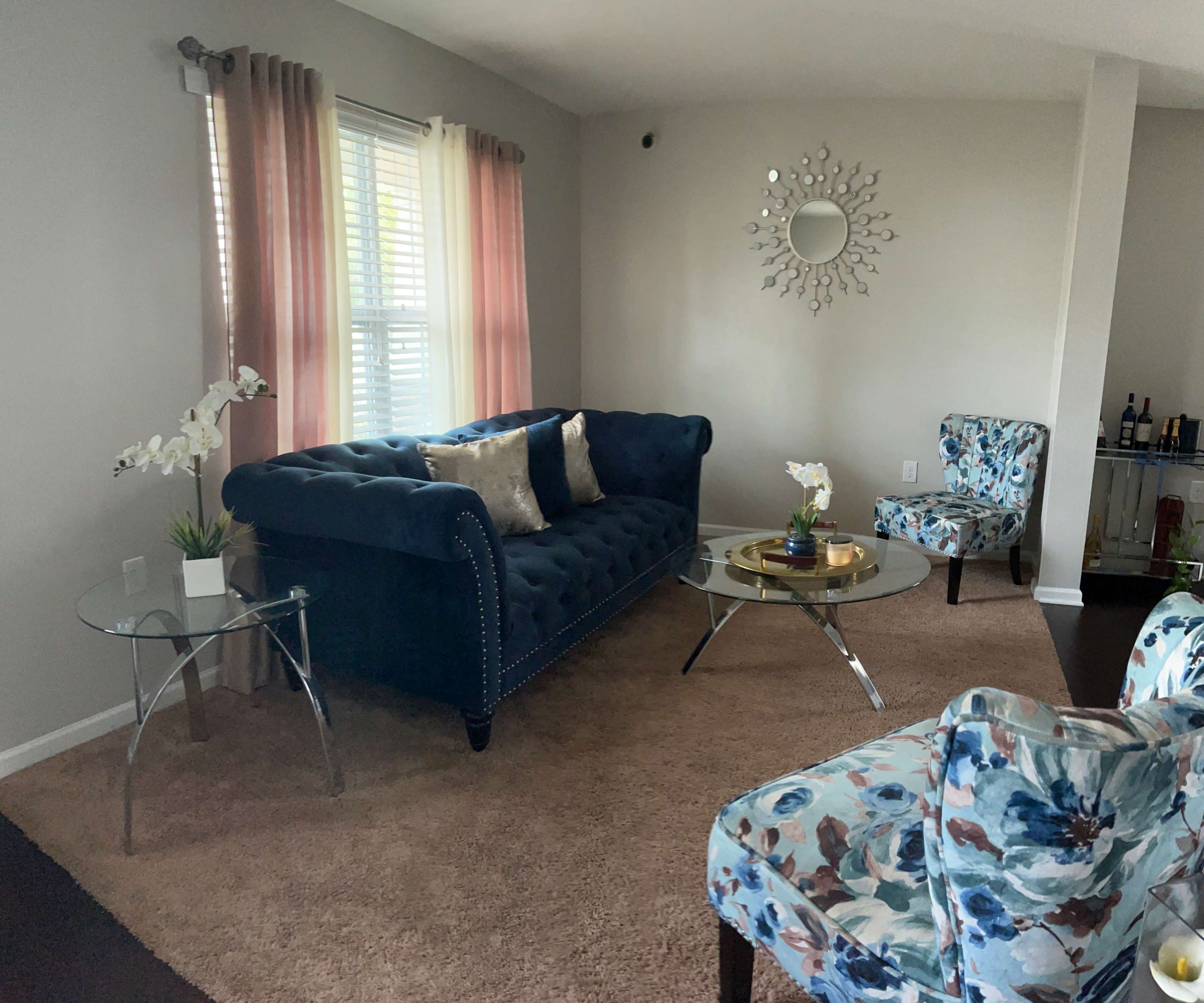 The living room features a blue tufted sofa, two floral-patterned chairs, a glass coffee table, and large windows with light curtains.