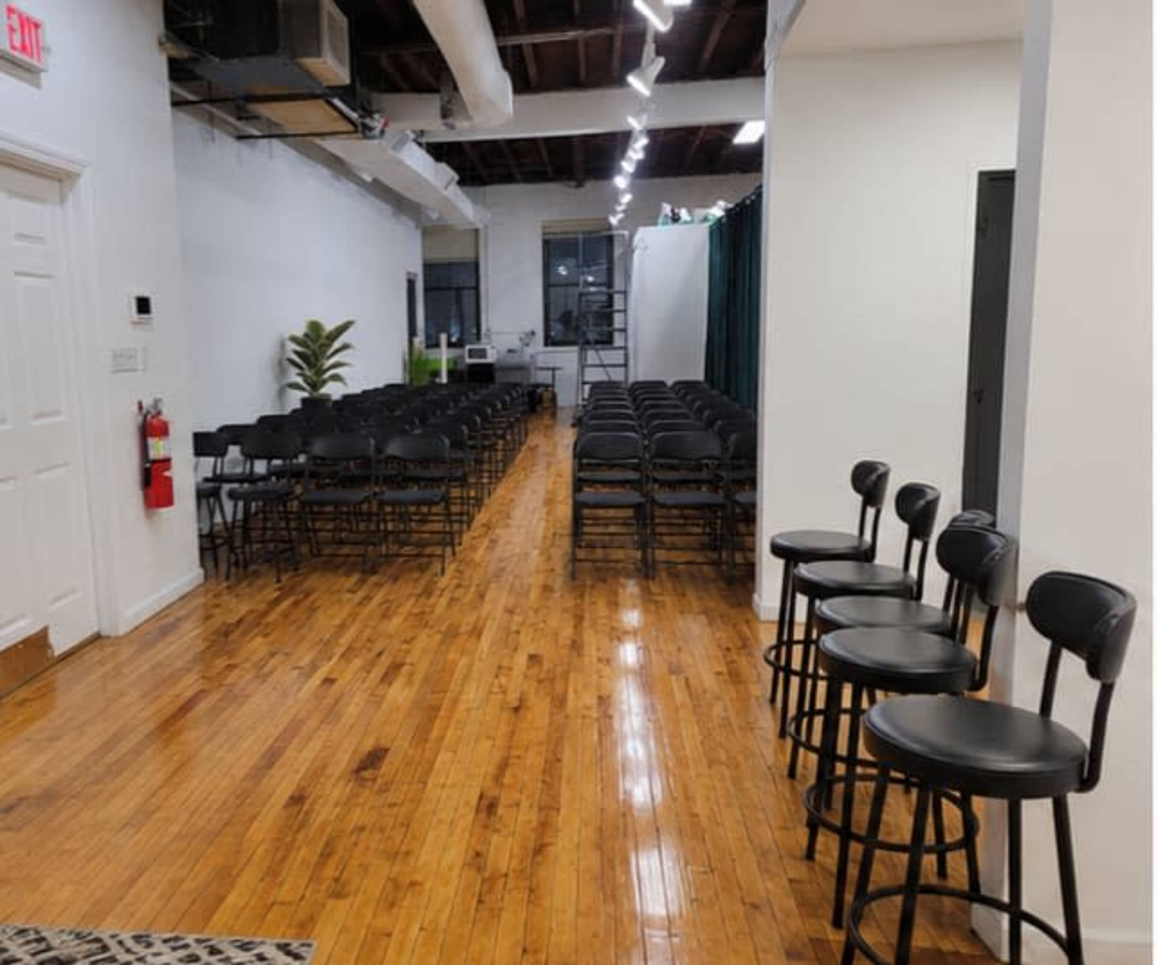 The image shows a spacious interior with two rows of black folding chairs arranged for an event, alongside black bar stools against one wall.