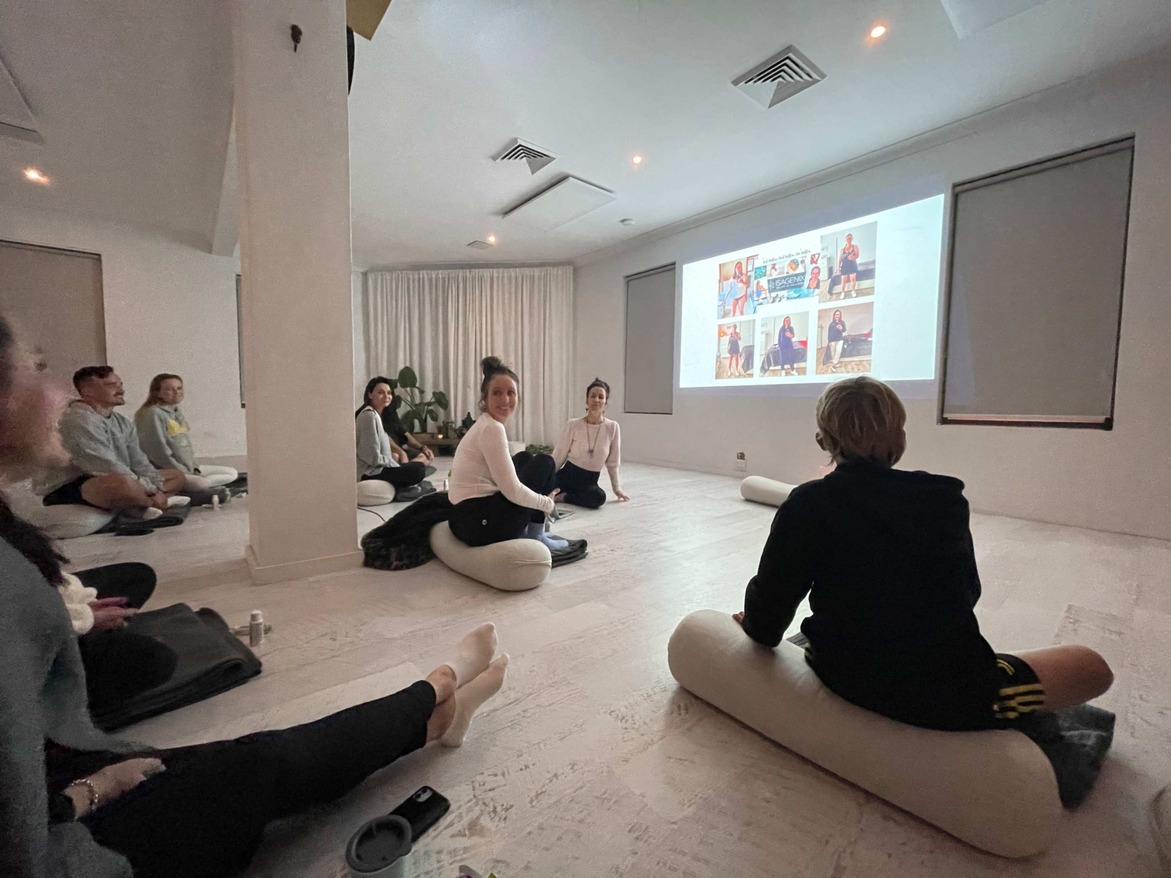 A group of individuals sits on floor cushions in a bright room while watching a presentation on a screen.