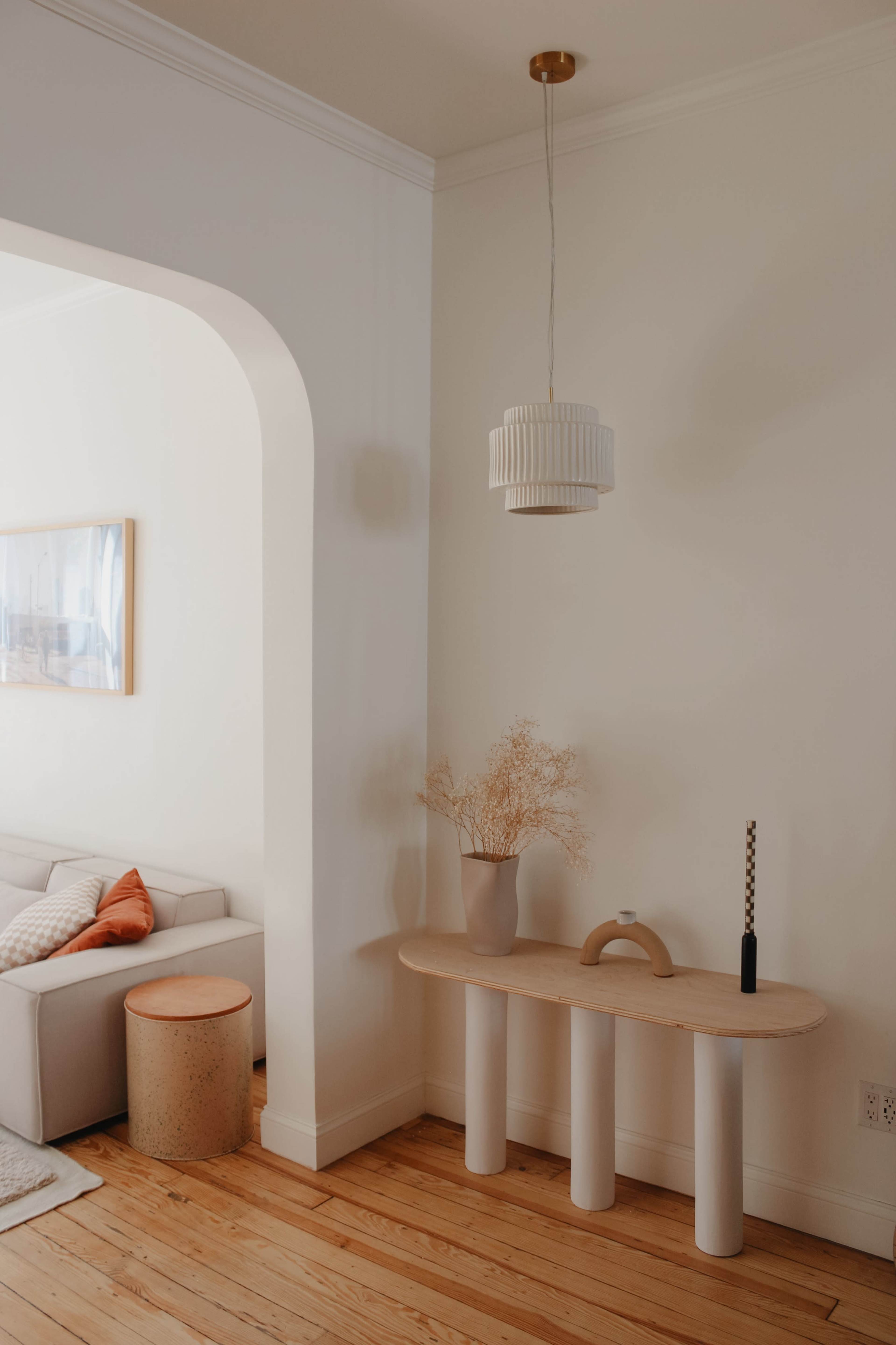 A minimalist living room features a light wooden console table with a vase of dried flowers, a lamp, and a cylindrical stool beside a beige sofa.