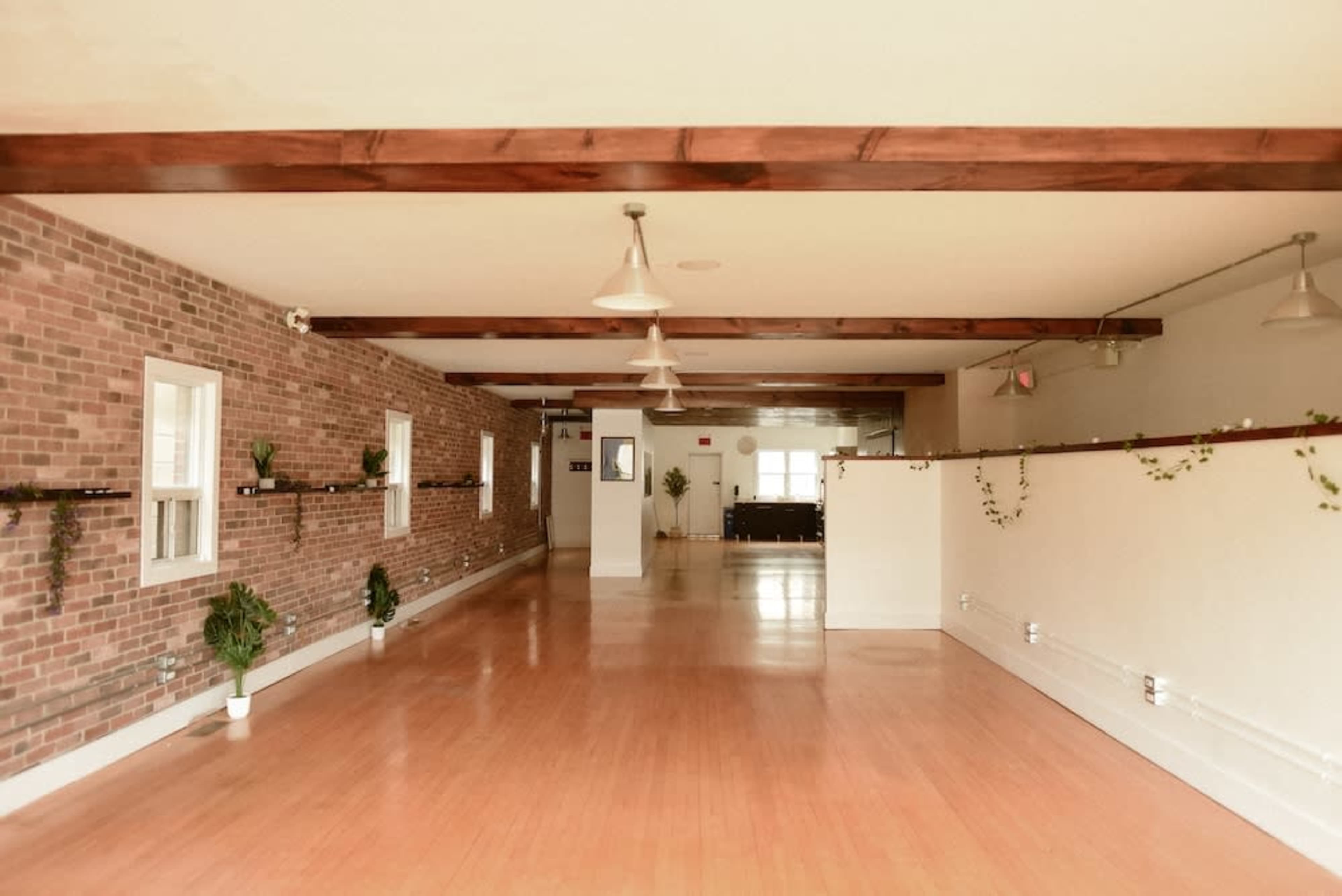 The image shows a spacious interior room with wooden beams, exposed brick walls, and a polished wooden floor, featuring plants along the walls.