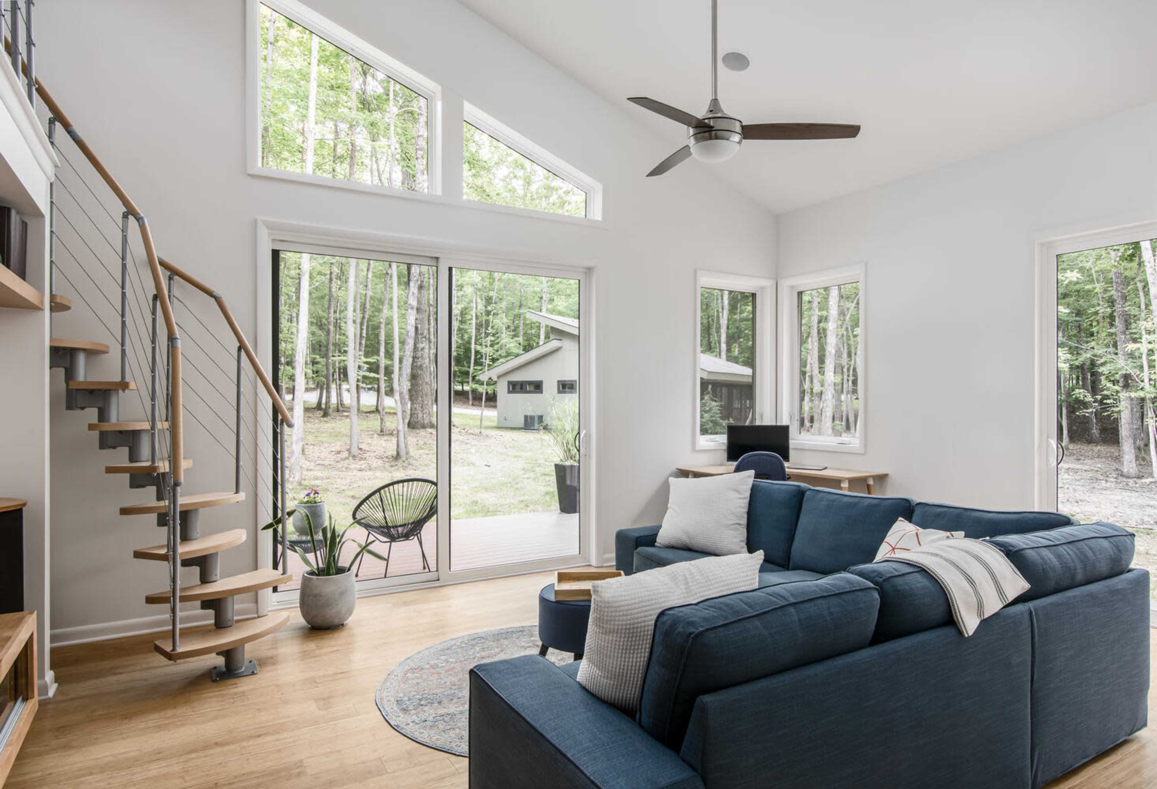 A modern living room features a blue sectional sofa, large windows overlooking a wooded area, and a spiral staircase leading to an upper level.