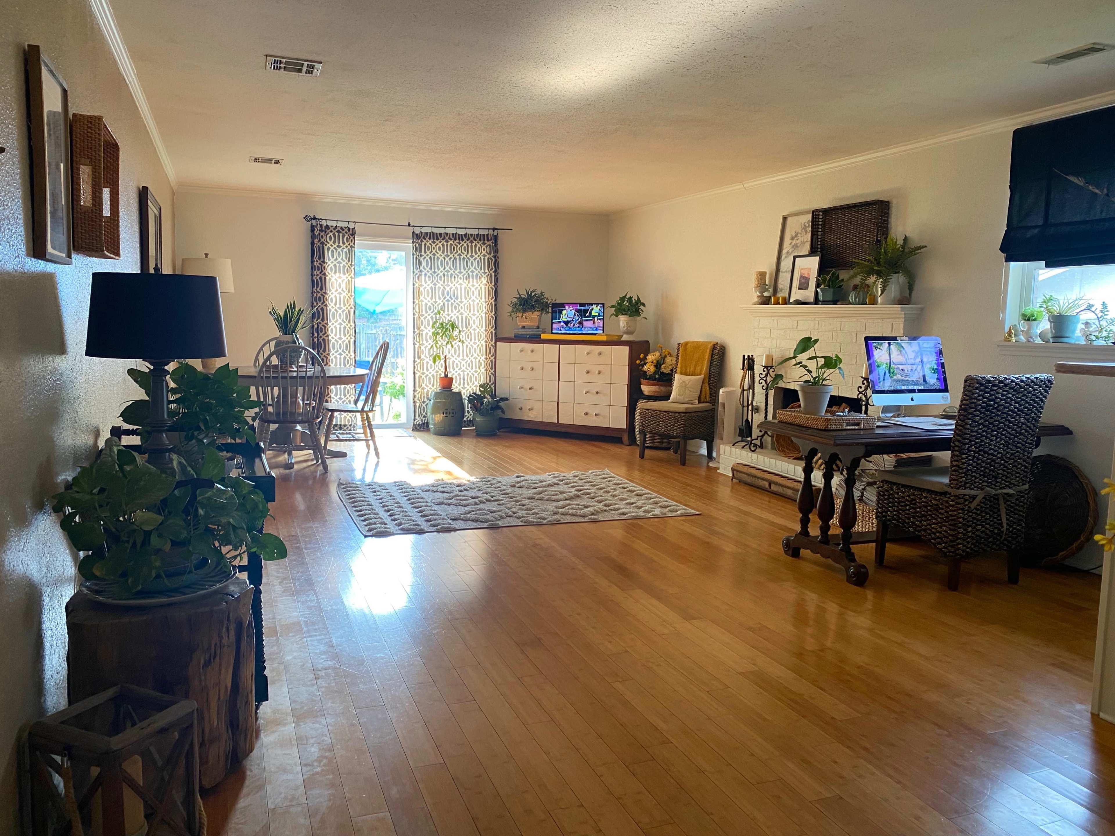 The image shows a spacious living room with hardwood floors, a dining area, a workspace with a desk, and various potted plants scattered throughout.