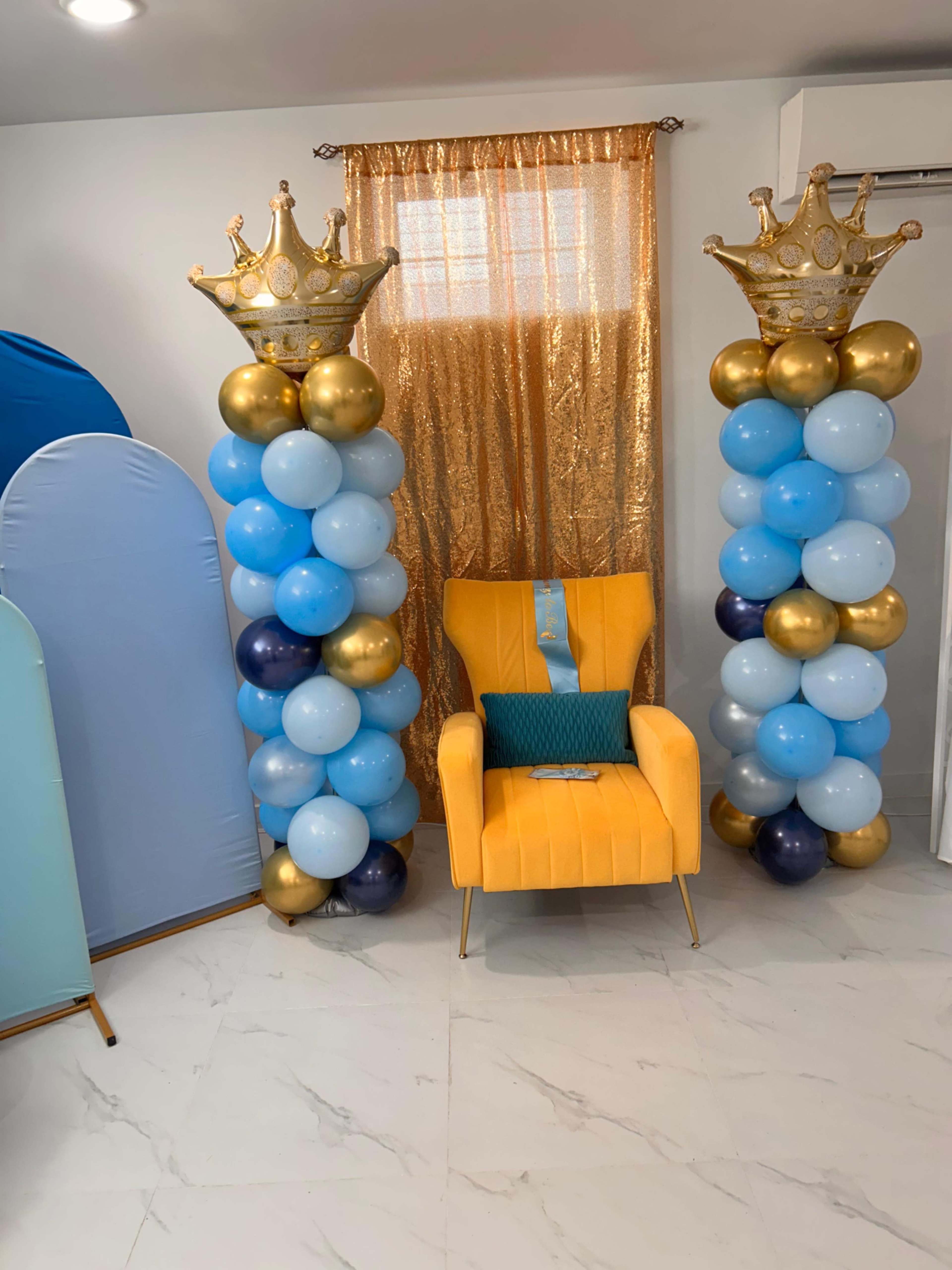 The scene features a yellow armchair with a blue cushion positioned between two balloon columns adorned with crowns, set against a backdrop of a golden curtain.
