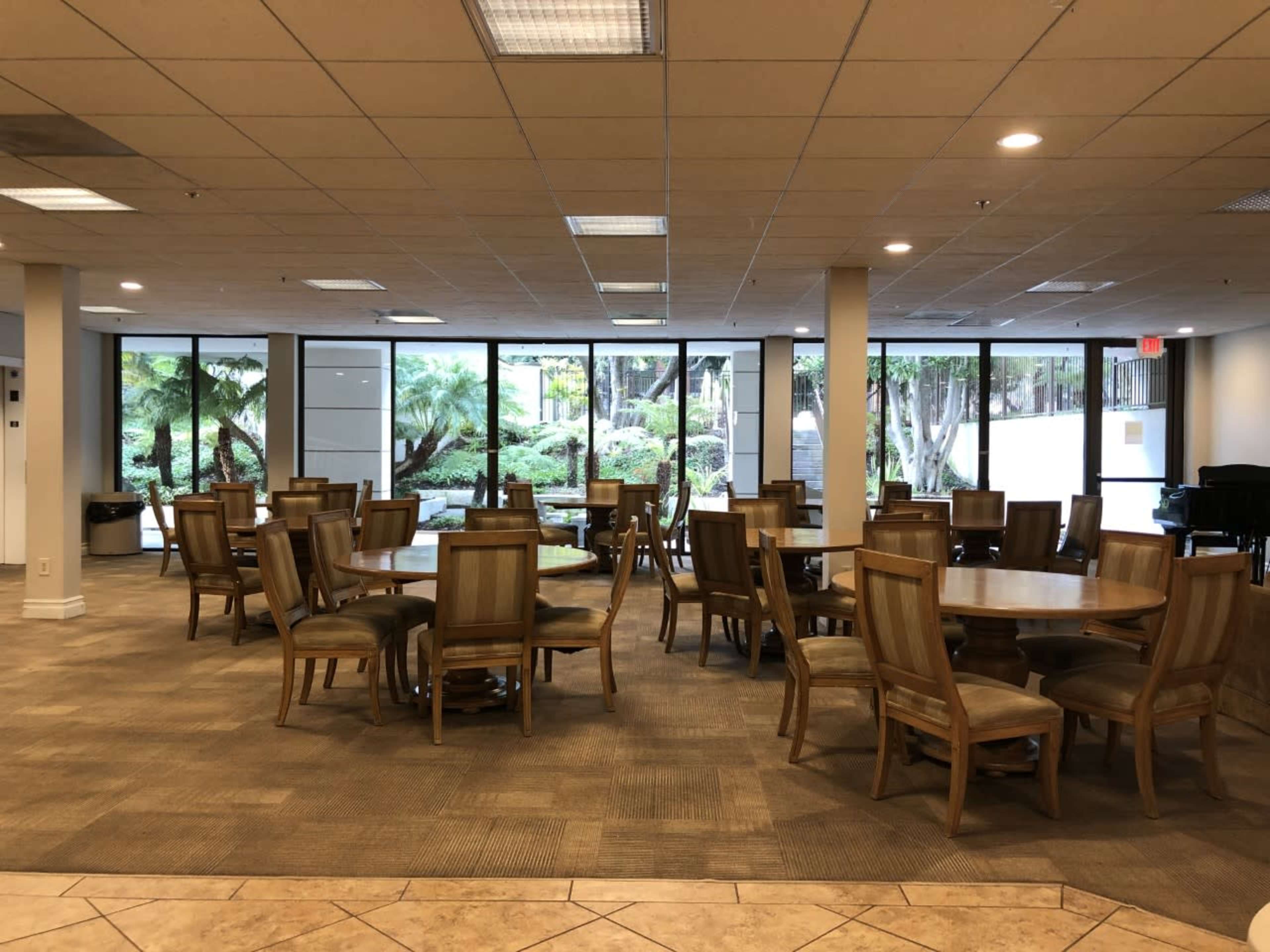 A spacious dining area with wooden tables and chairs arranged in rows, surrounded by large windows that overlook a green outdoor landscape.