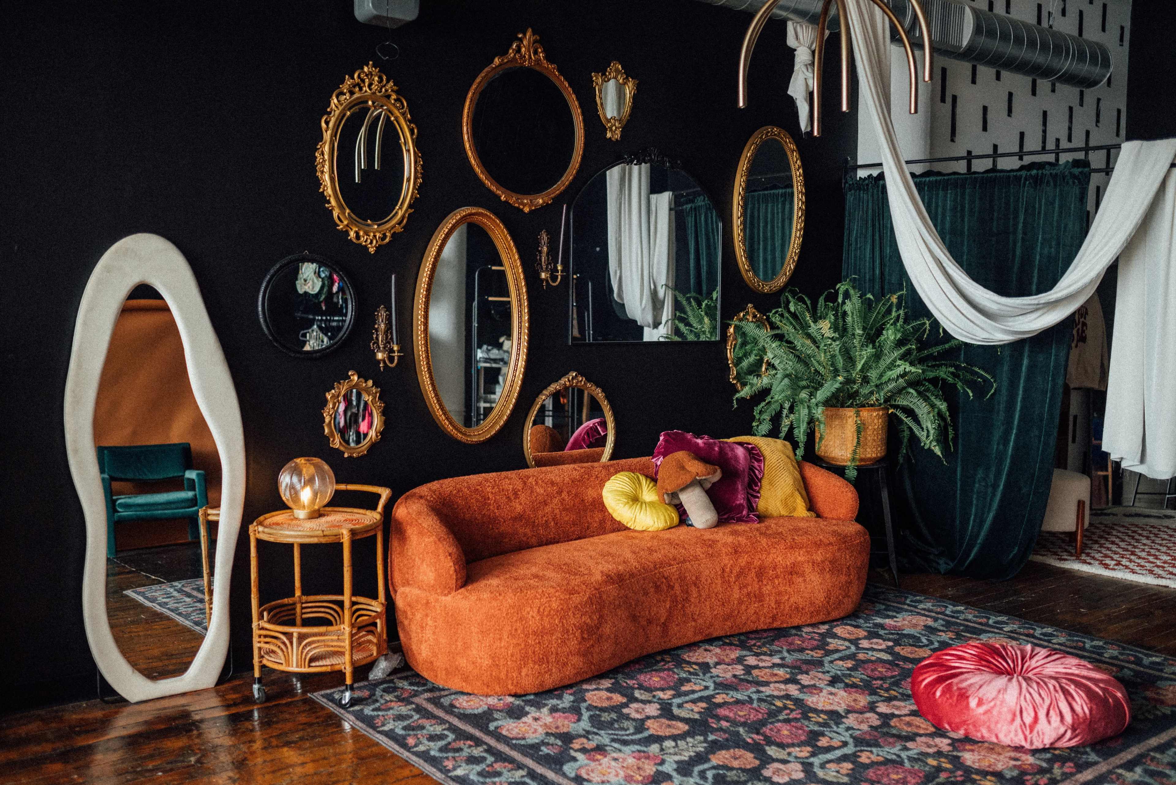 A stylish living room features a curved orange sofa, decorative mirrors on the black wall, and a colorful rug with a mix of plants and accessories.