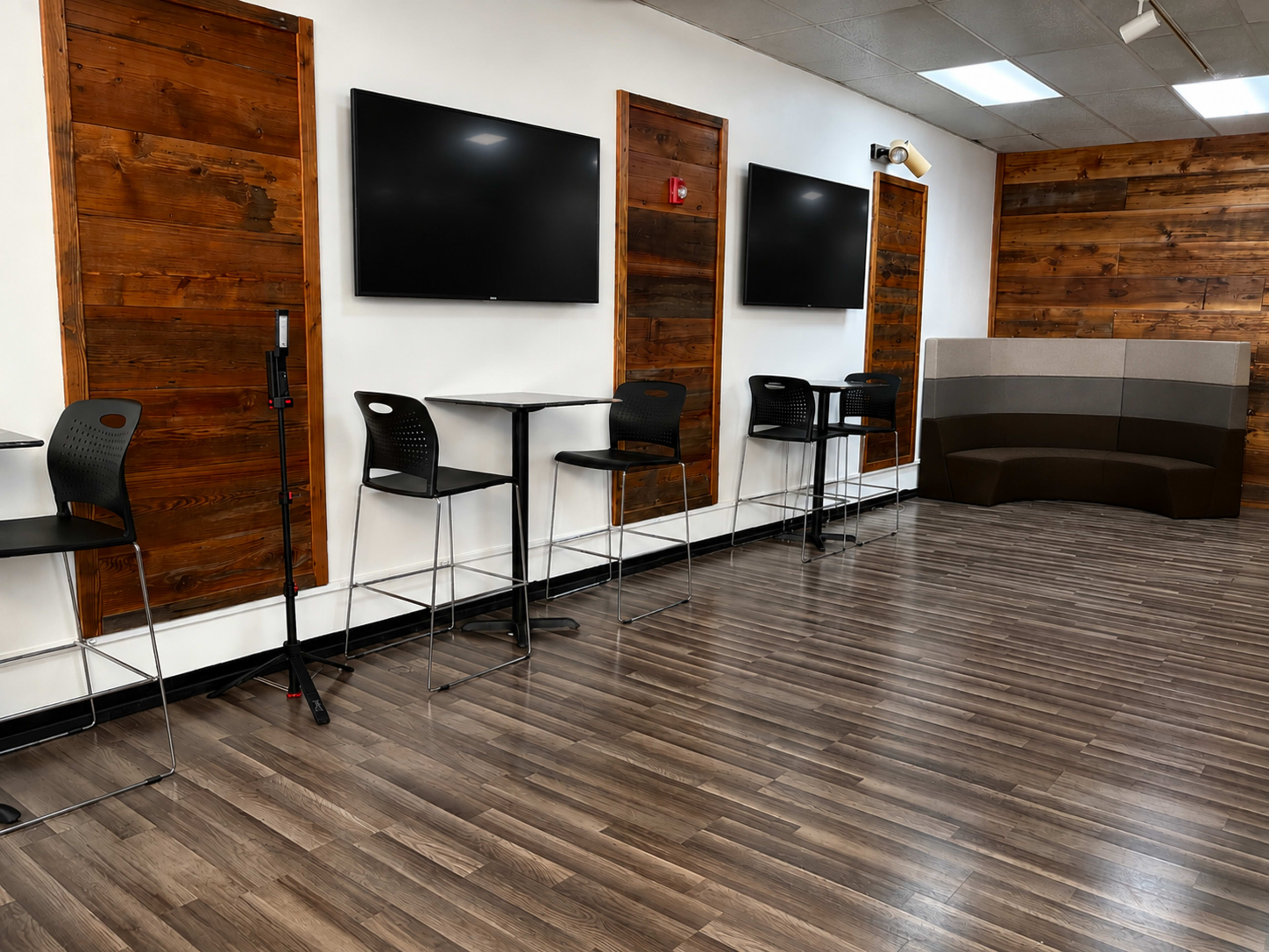 The image shows a spacious room with wooden paneling, two wall-mounted televisions, and black high-top tables with chairs arranged along one side.