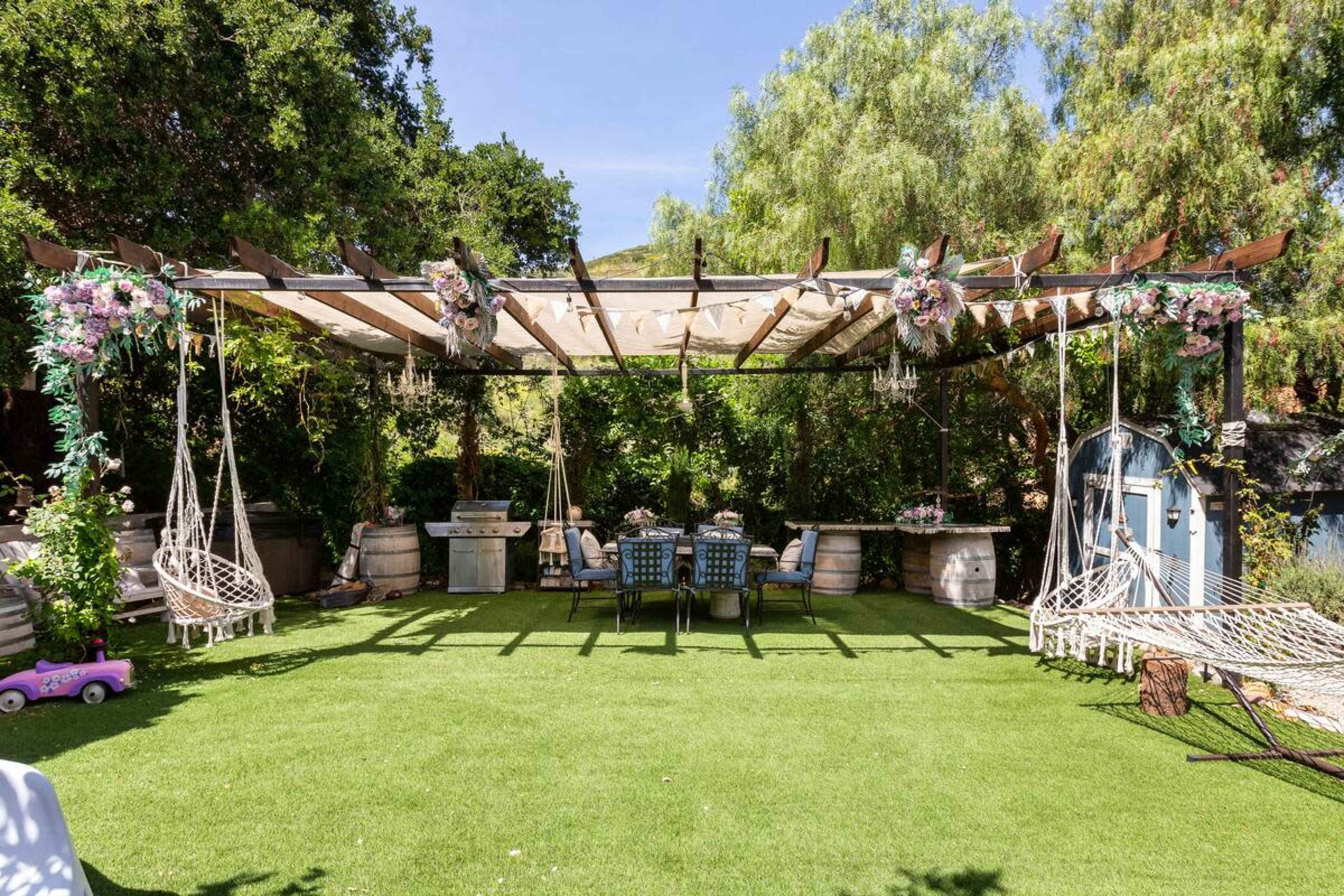 The image depicts a backyard patio area with a wooden pergola, adorned with floral decorations, featuring a table and chairs, swinging hammocks, and green grass.