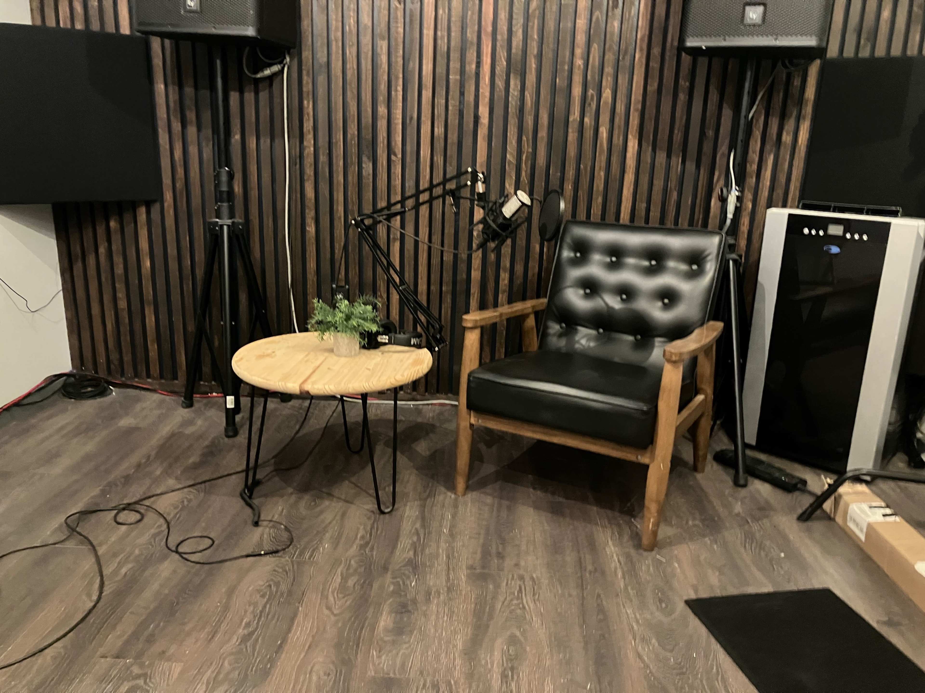The image shows a small recording studio with a black leather chair, a wooden table, a microphone on a stand, and audio equipment against a wooden panel backdrop.