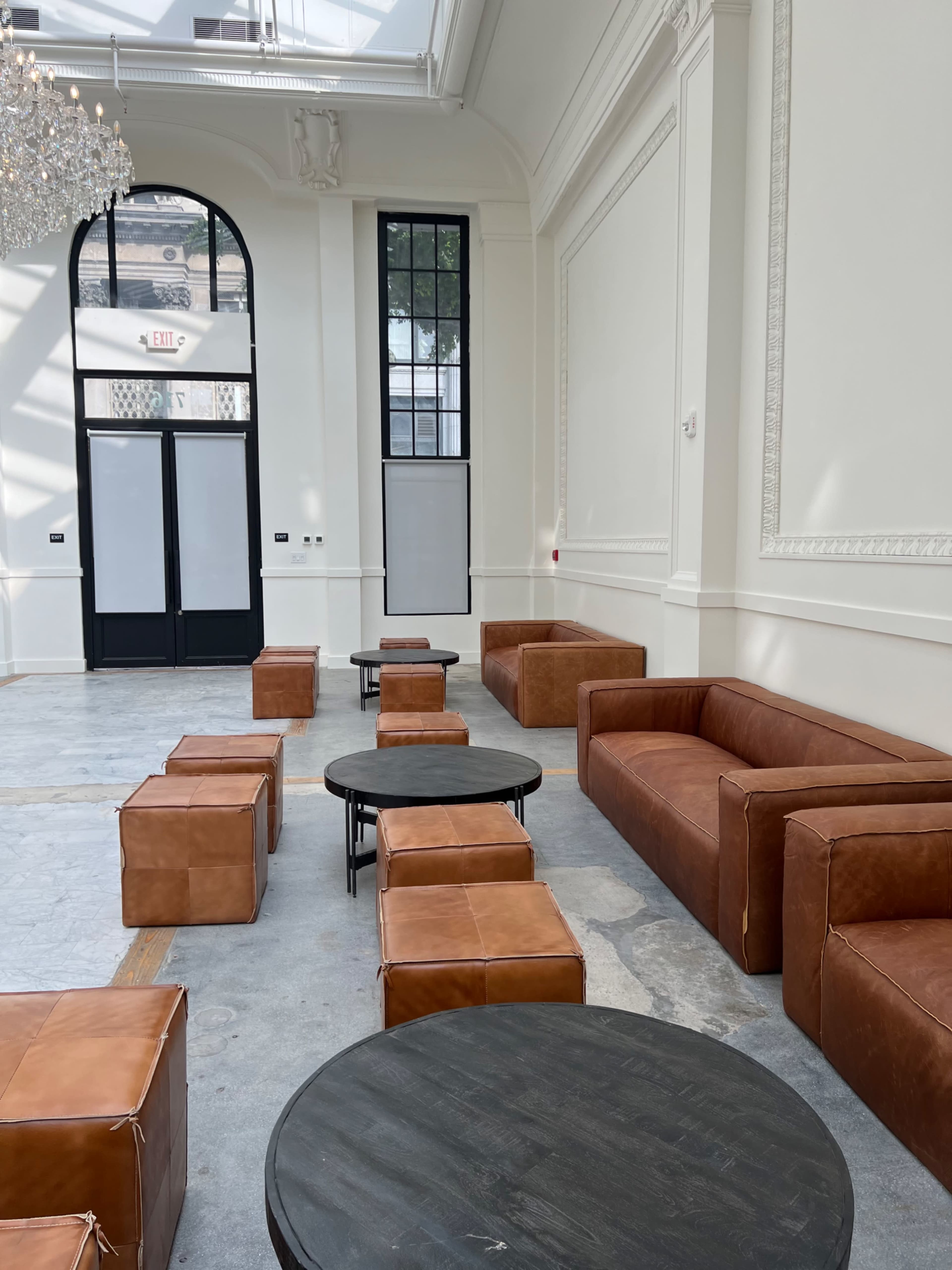 The image shows a spacious room with high ceilings, featuring brown leather sofas and stools arranged around dark round tables.