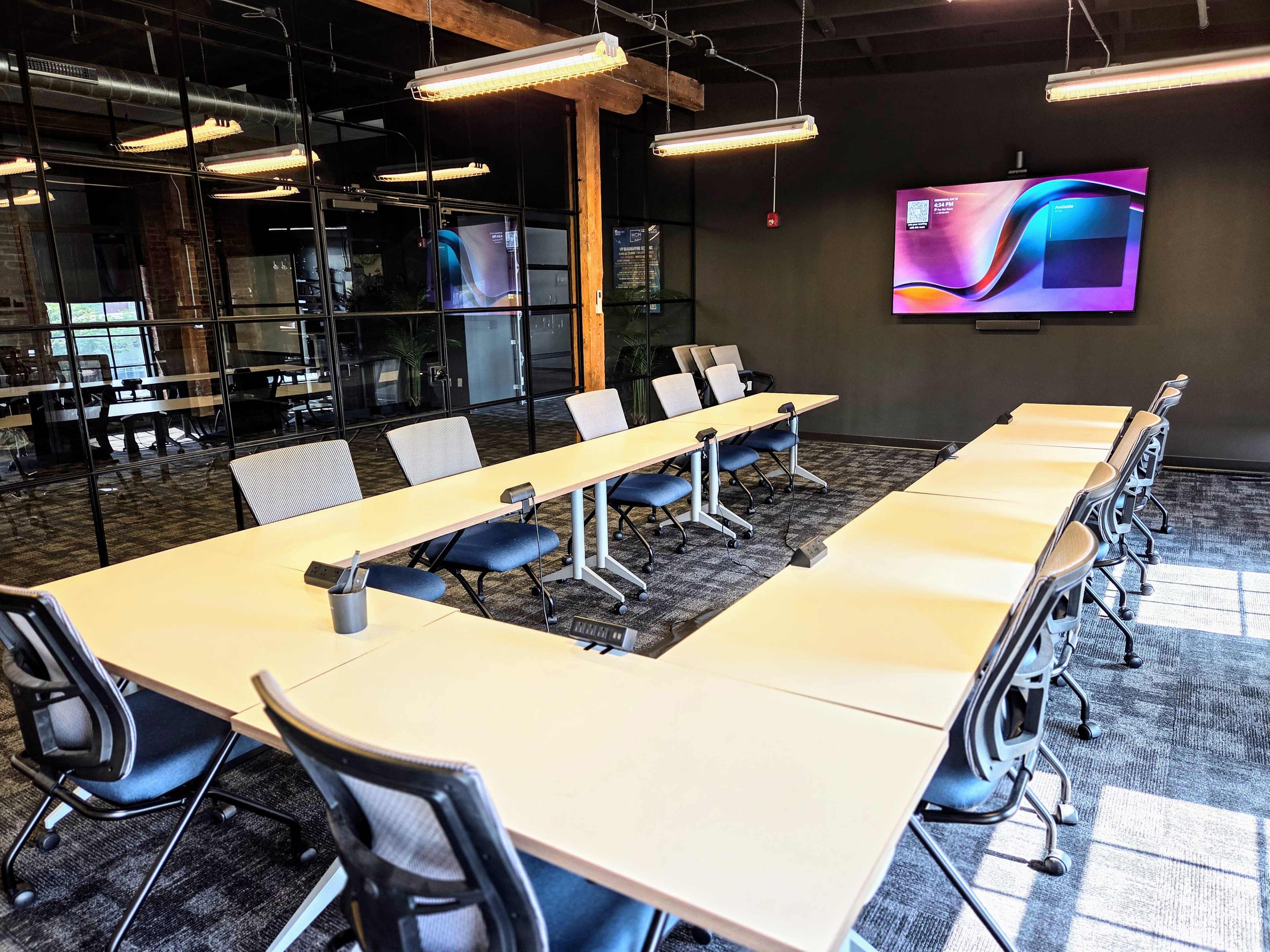 A modern conference room features a long table surrounded by ergonomic chairs, with a large screen mounted on the wall and glass partitions in the background.
