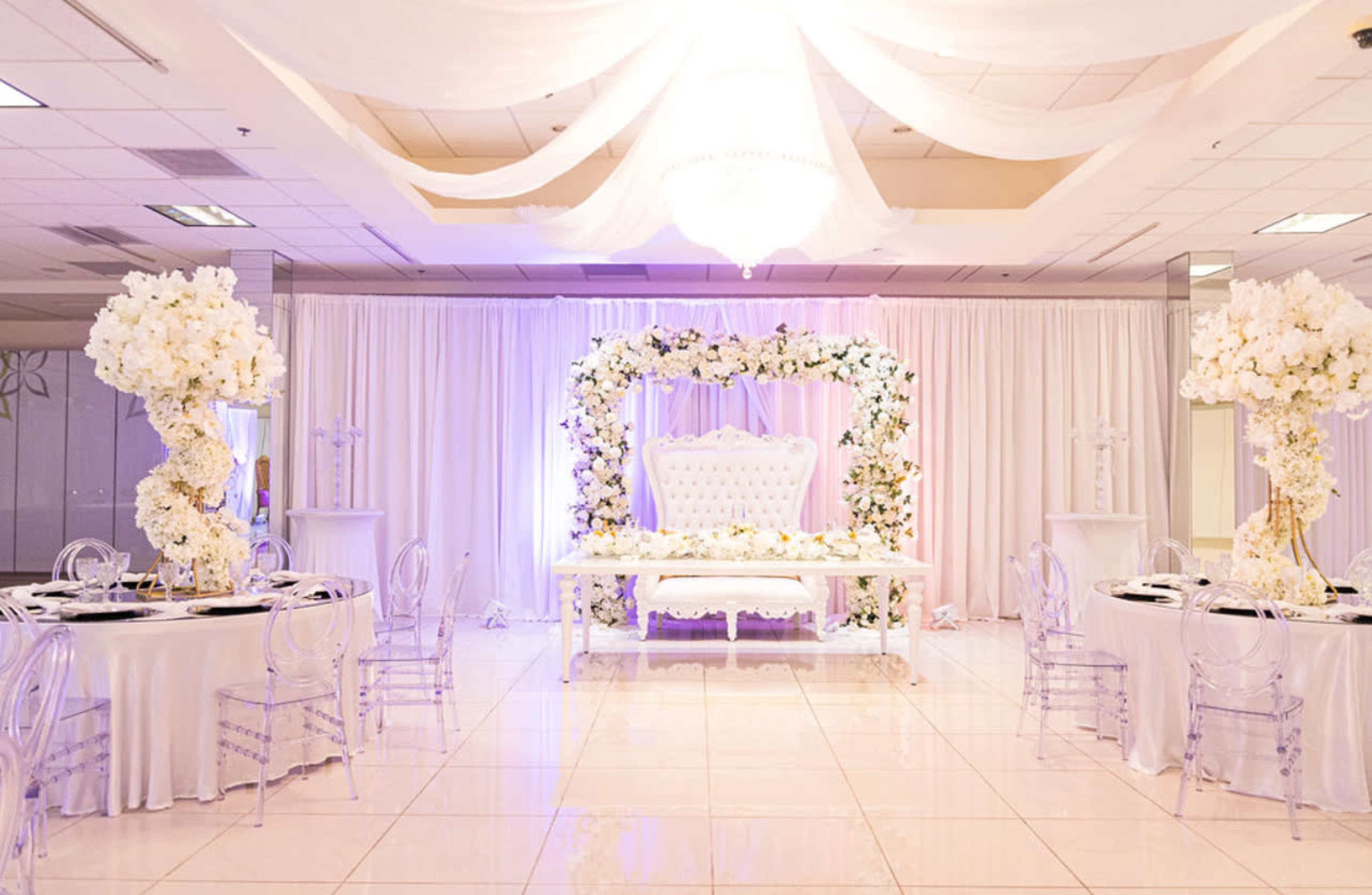 An elegantly decorated event space featuring a white floral arch, a couple's seating area, and round tables set with white linens and clear chairs.
