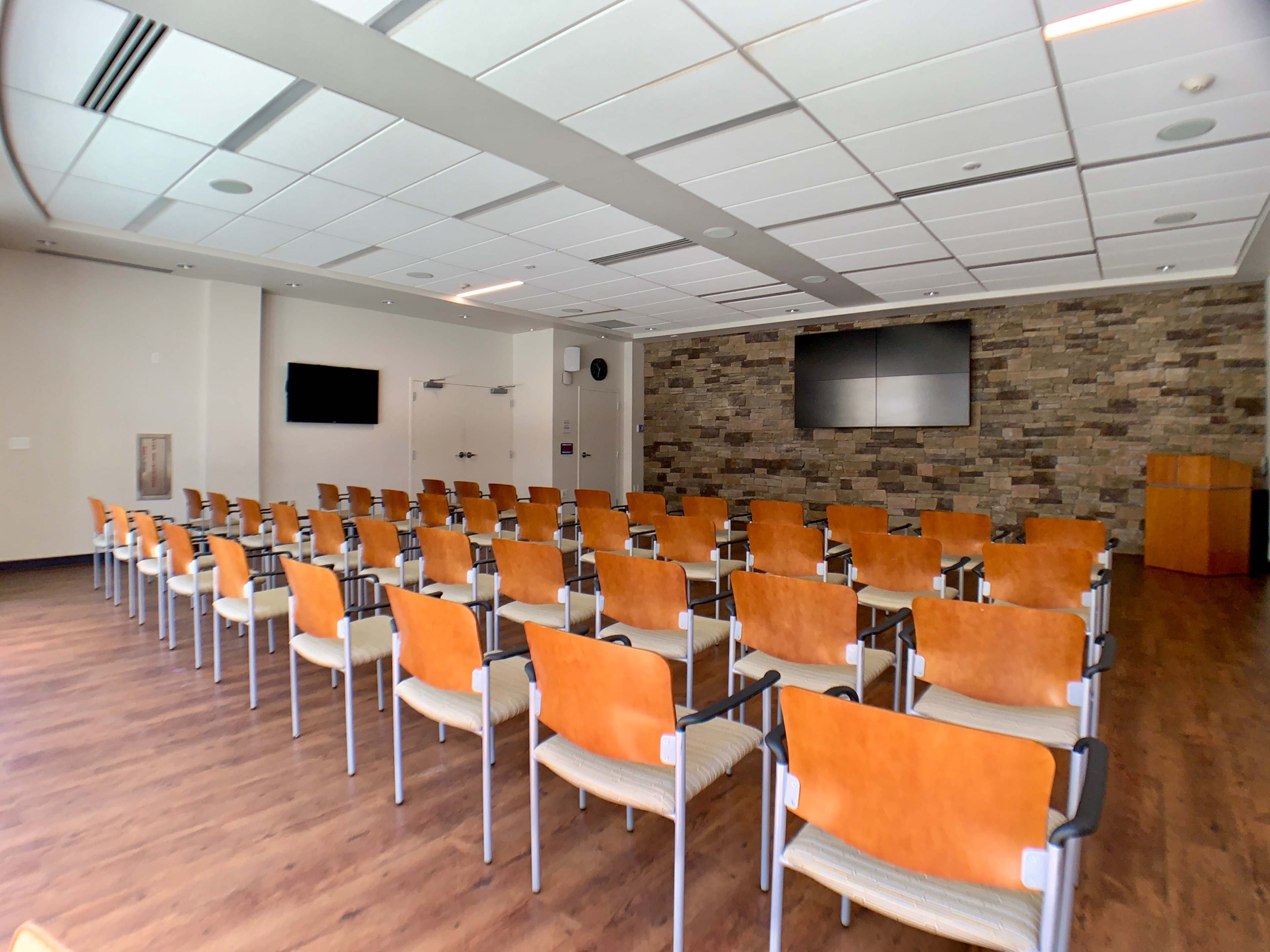 A conference room with rows of orange and white chairs facing a stone wall featuring two large screens and a wooden podium.