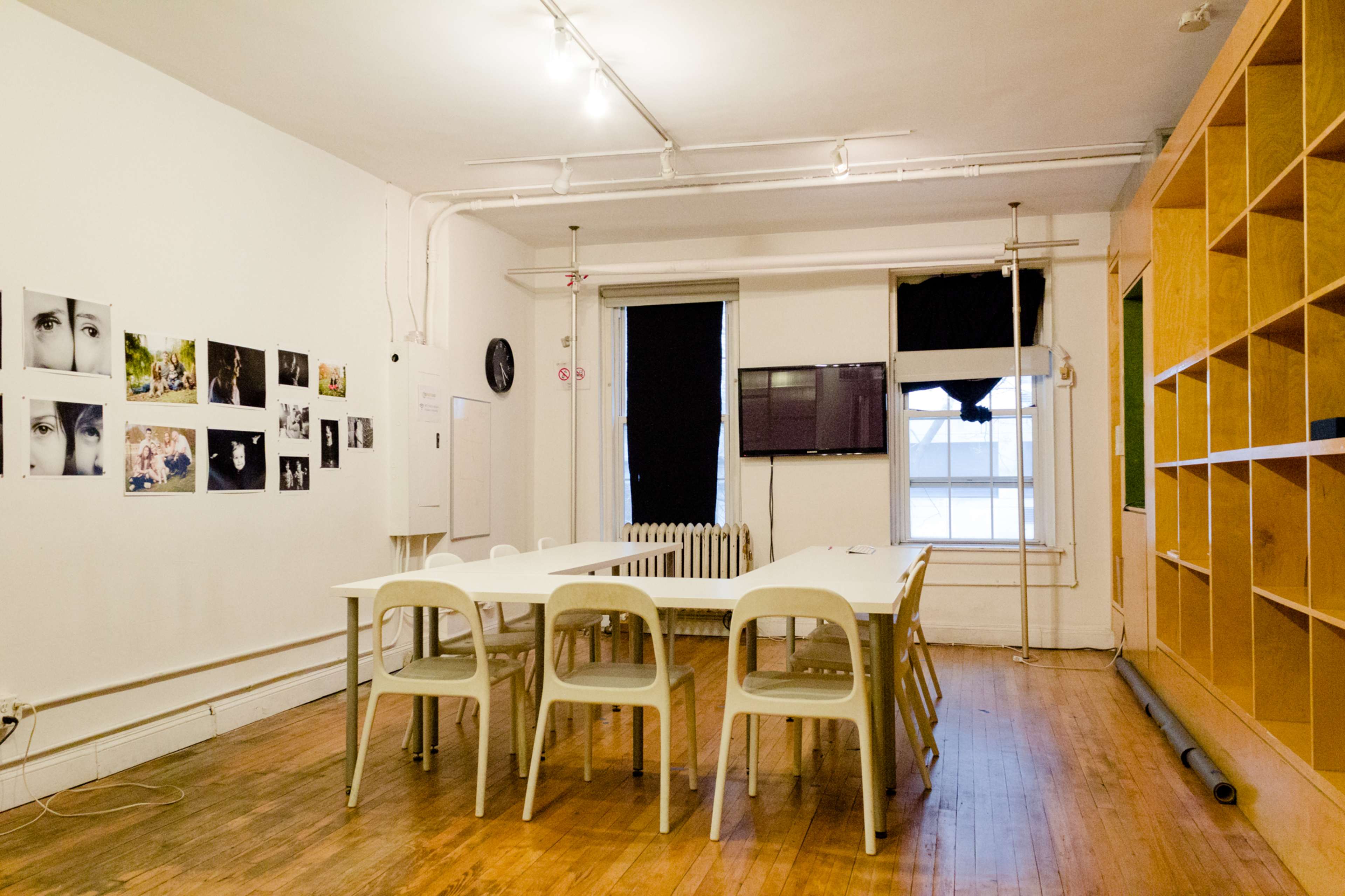 A spacious room features a long table surrounded by chairs, with photographs on the wall, a black television, and wooden shelving.