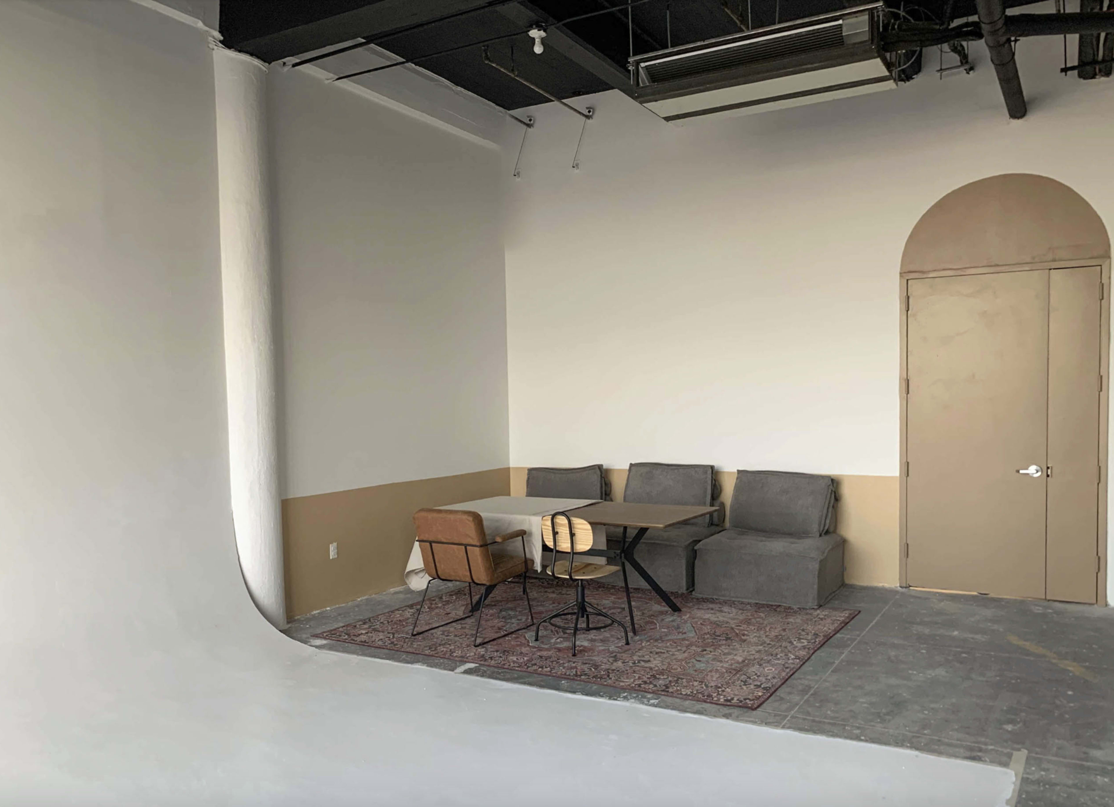 The image shows a minimalist studio space featuring a simple seating area with gray couches and a wooden table surrounded by chairs, against a backdrop of plain walls.