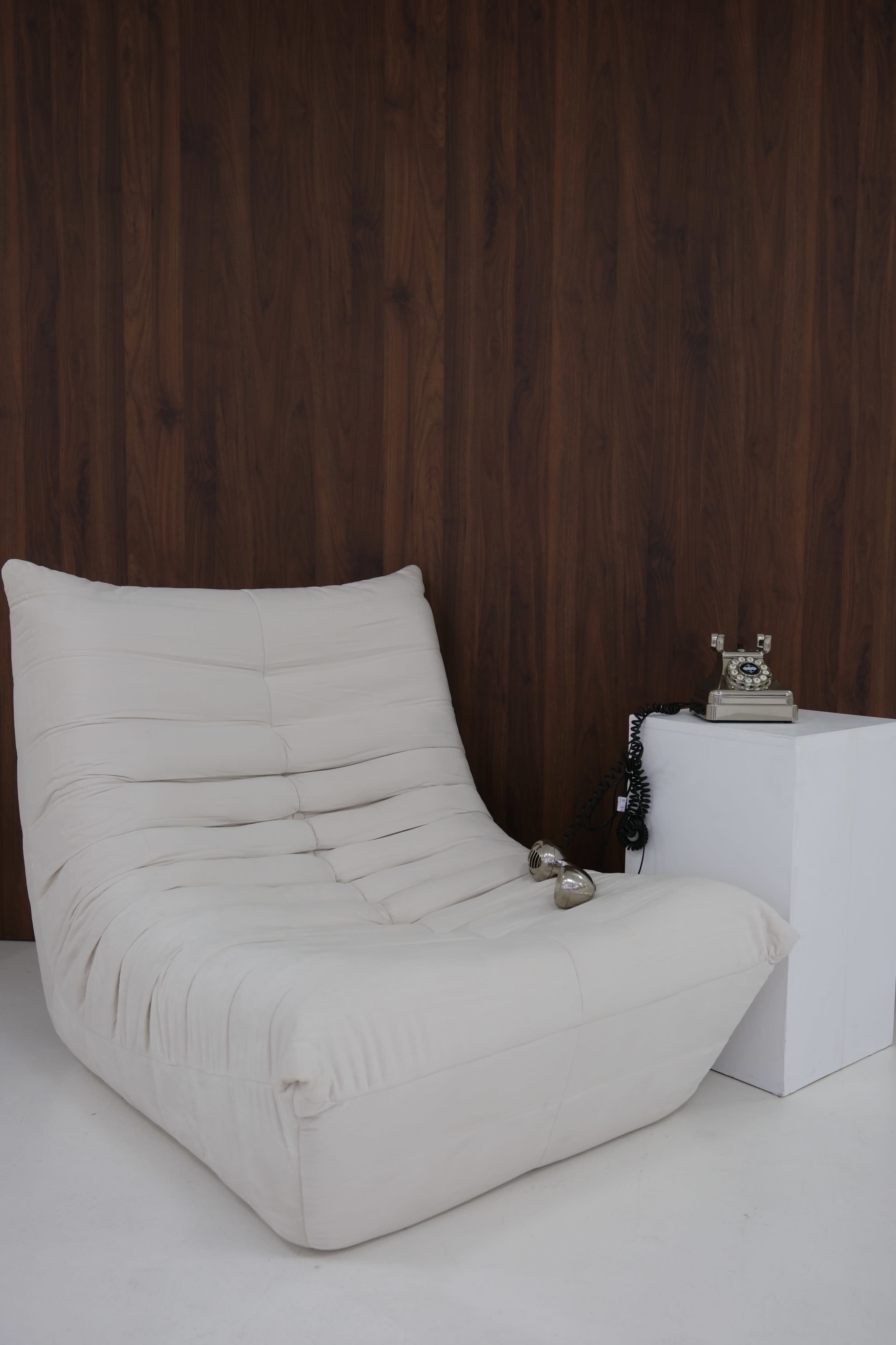The image shows a modern, white lounge chair next to a small white side table with a vintage rotary phone on it, against a wooden wall.