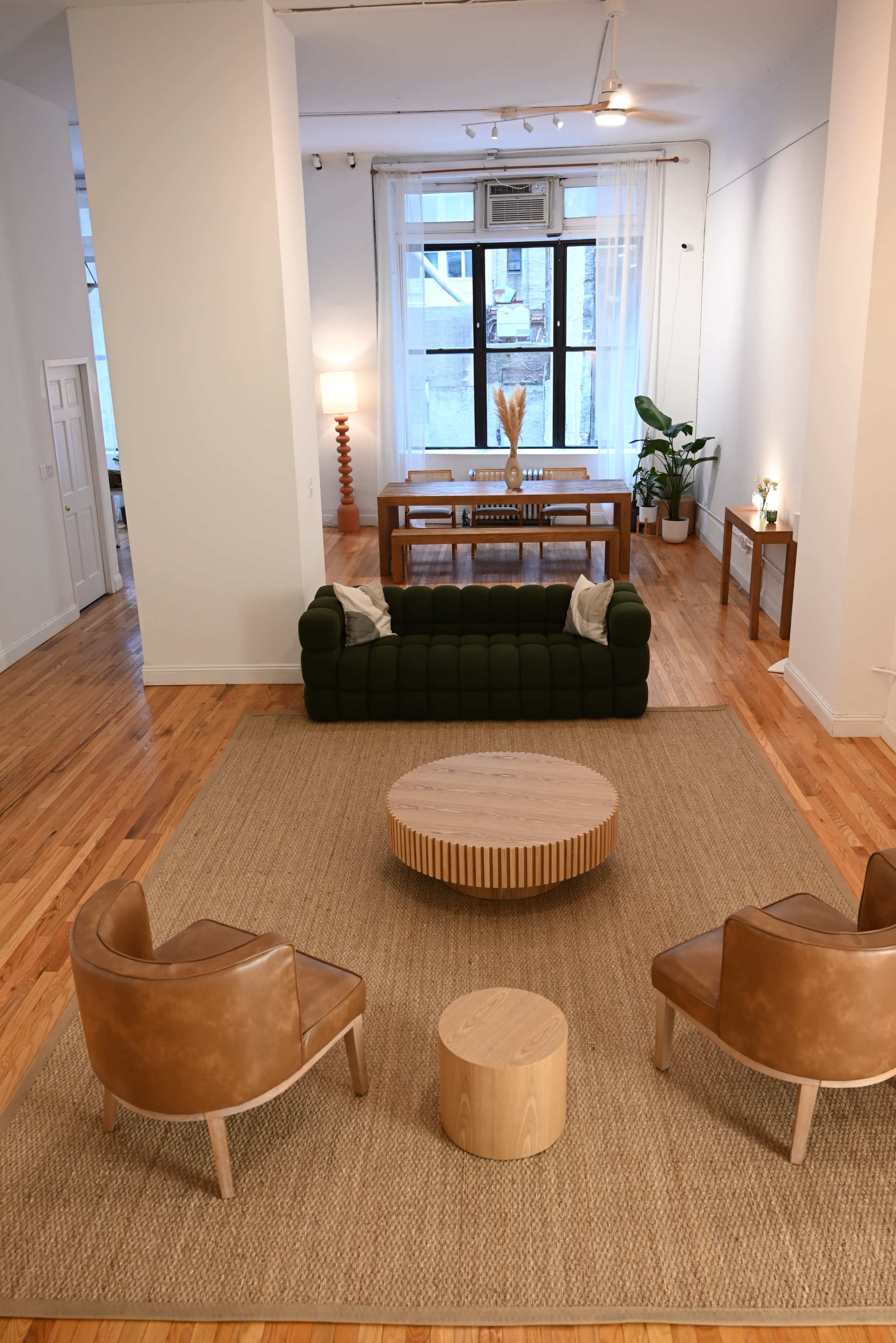 The image shows a spacious interior featuring a green couch, two brown chairs, a round coffee table, and a dining area in the background.