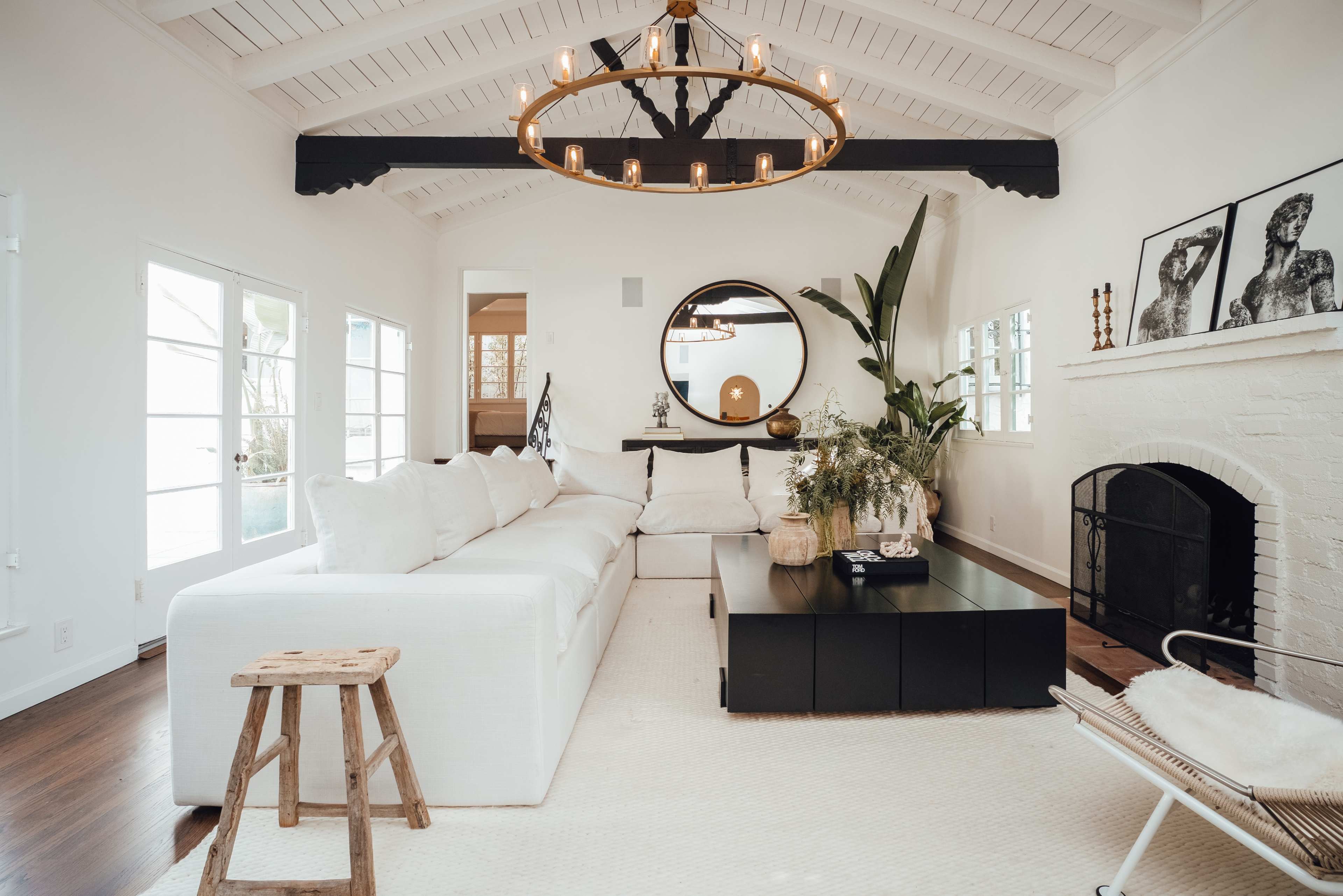 A spacious living room features a large white sectional sofa, a round mirror, and a black coffee table, with a fireplace and large windows allowing natural light.