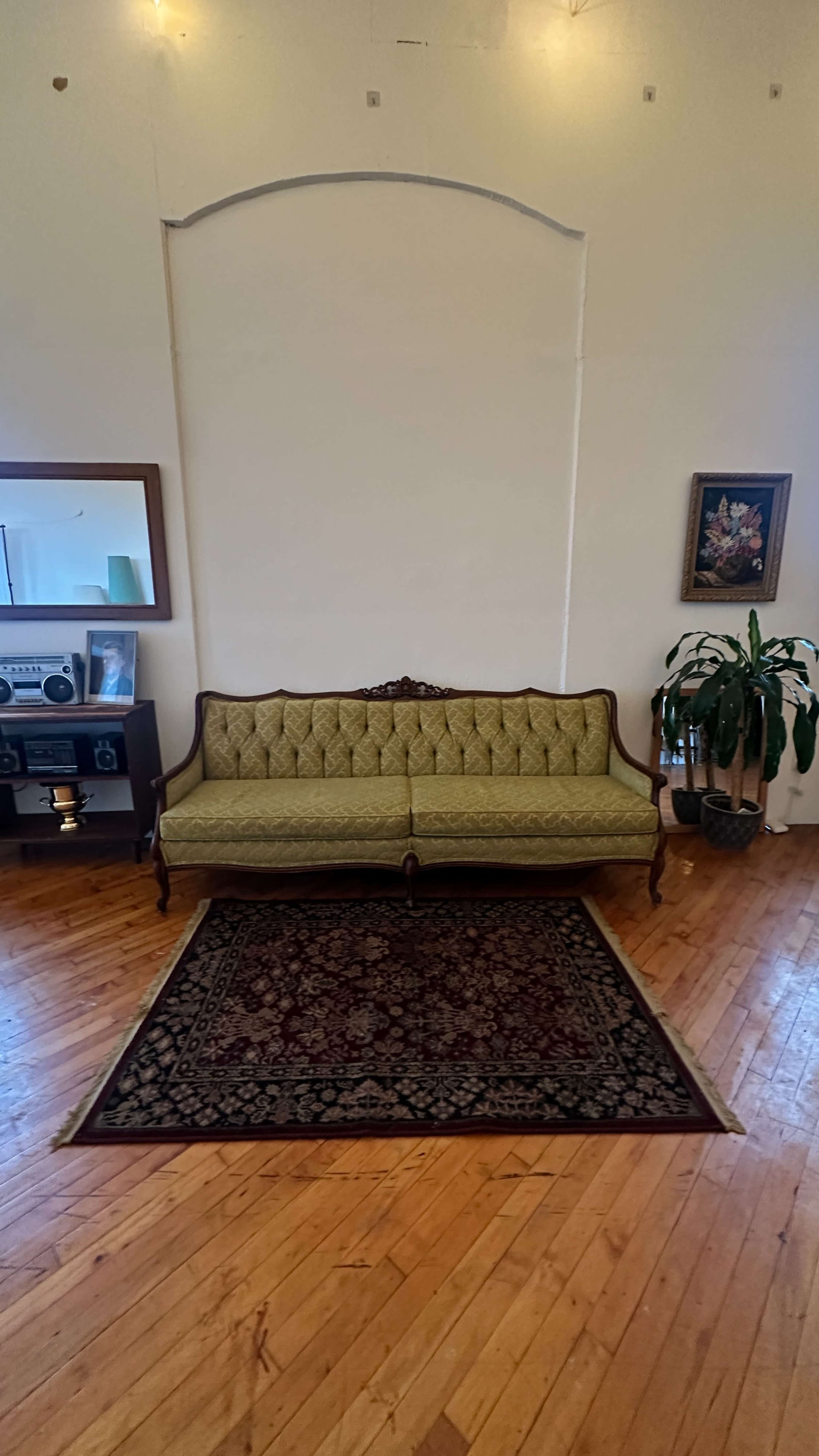 A green upholstered couch is positioned on a patterned rug, with a mirror and a small shelf displaying decorative objects nearby.