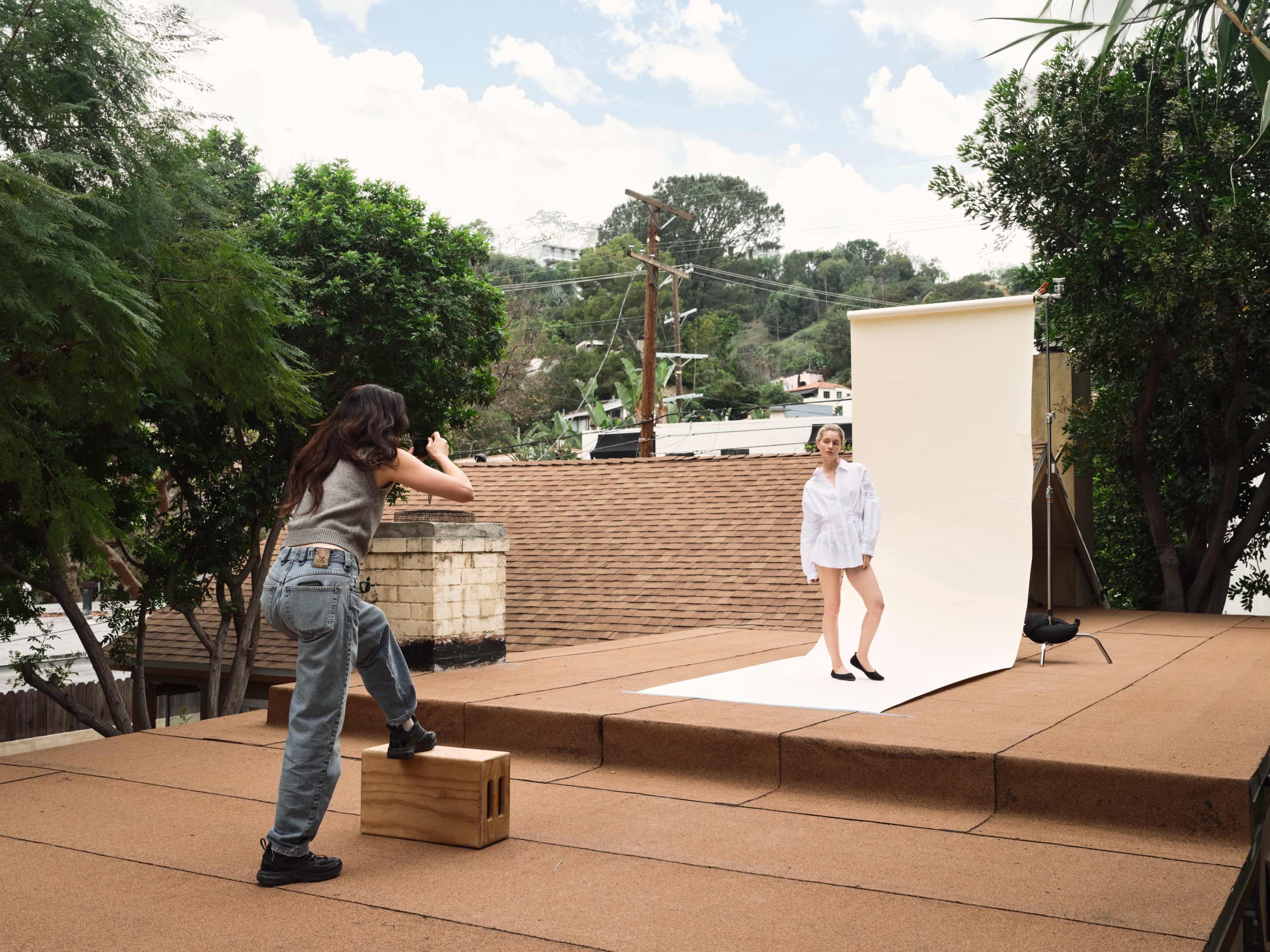 A person stands on a wooden crate, taking photos of a model posing in front of a white backdrop on a rooftop surrounded by greenery.