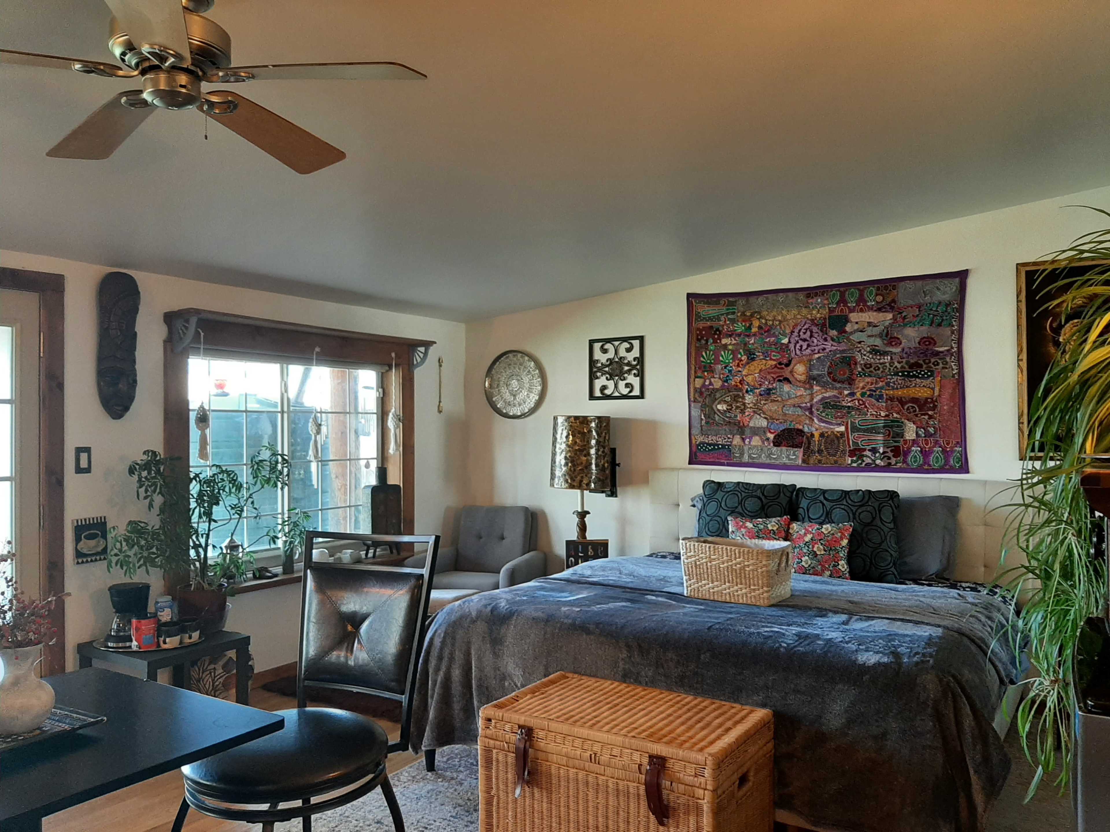 A cozy living space featuring a bed, armchair, decorative wall hangings, and various plants.