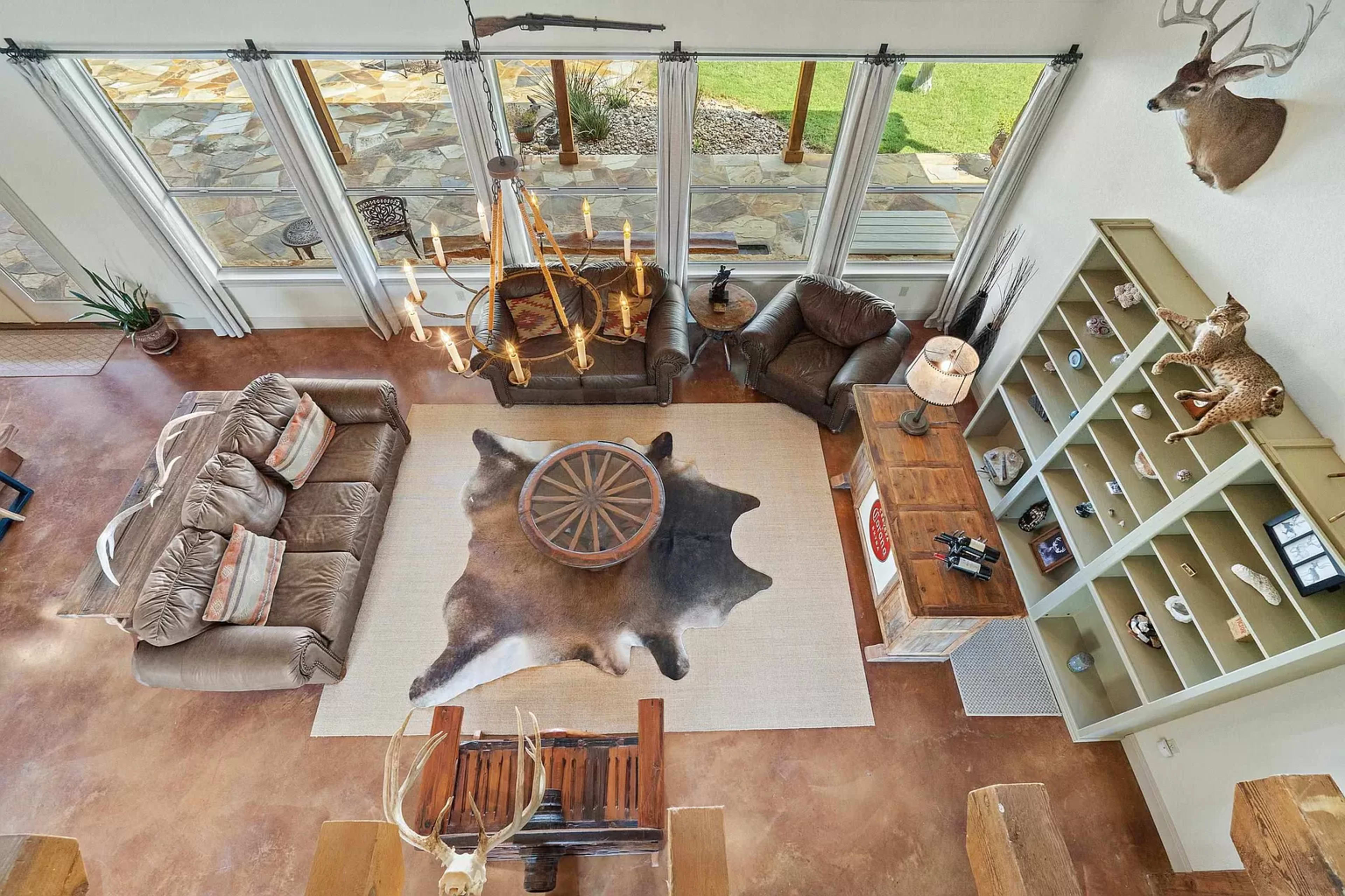 The image shows a spacious living room with large windows, a central area rug, a circular coffee table, and rustic decor, including mounted animal heads on the walls.