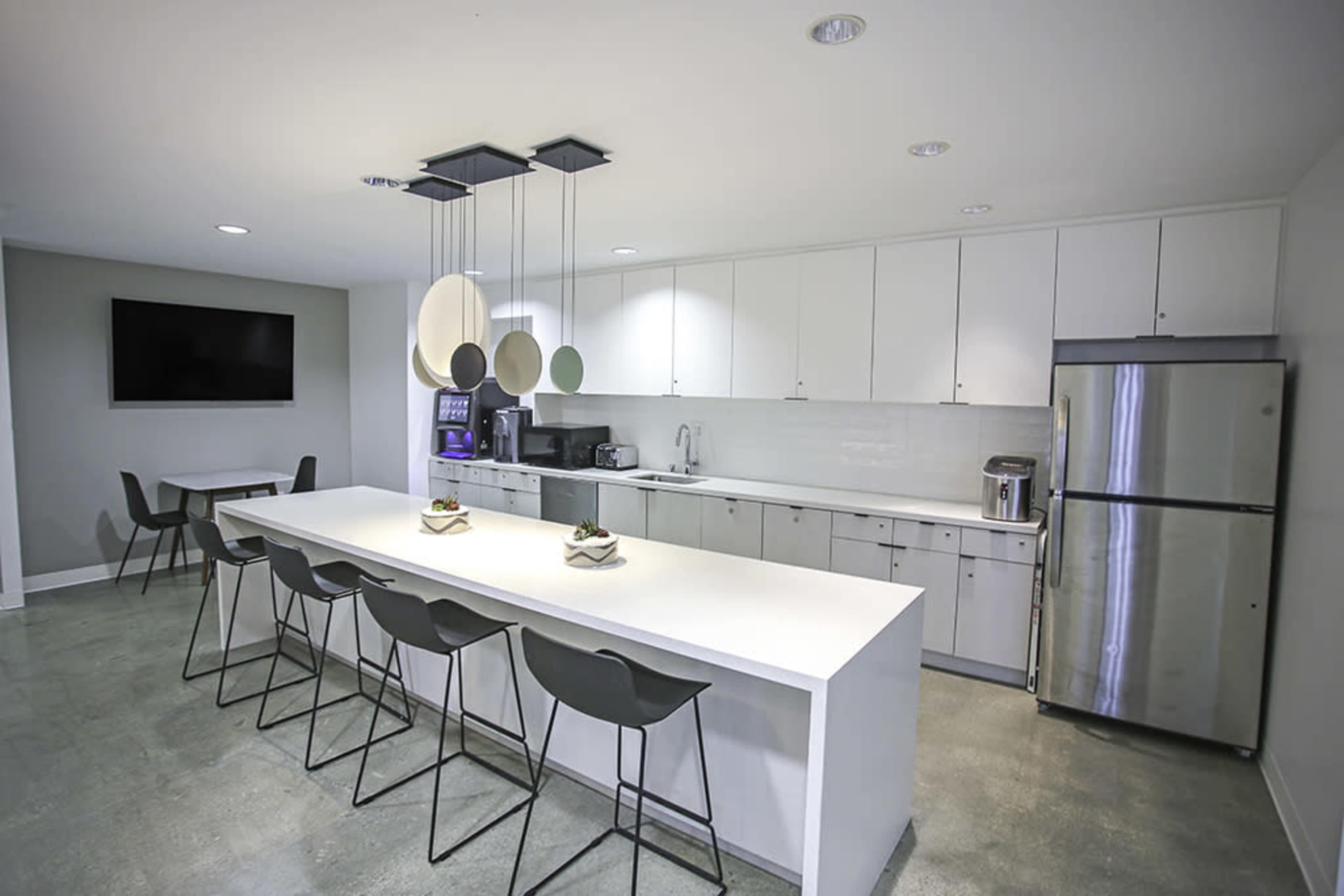 A modern kitchen equipped with a large white countertop, bar stools, stainless steel appliances, and a wall-mounted television.