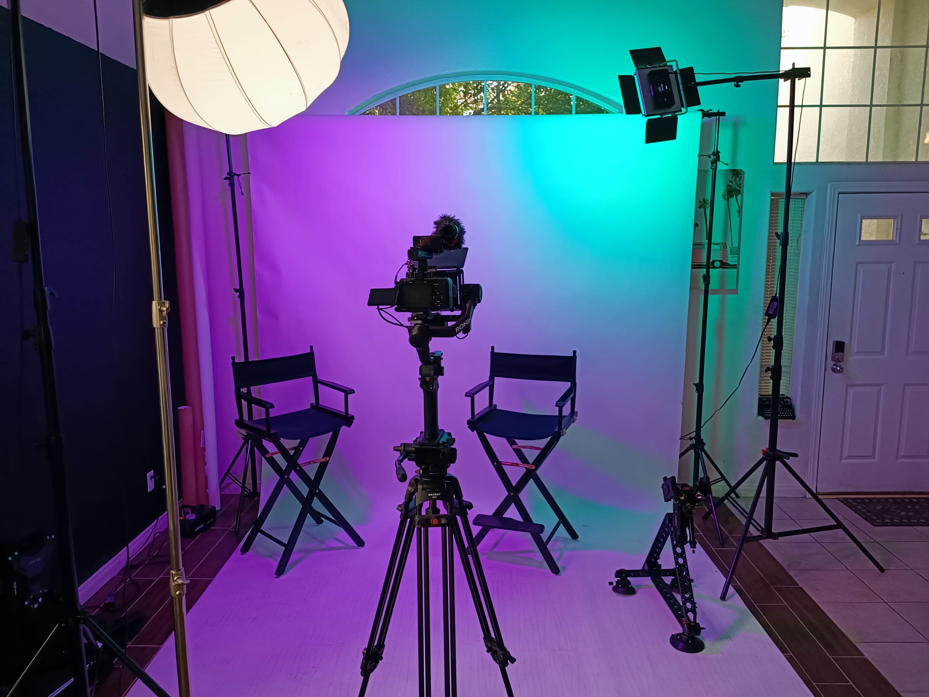 A professional photography setup with two director chairs, a camera on a tripod, and colorful lighting in a room with a front door visible in the background.