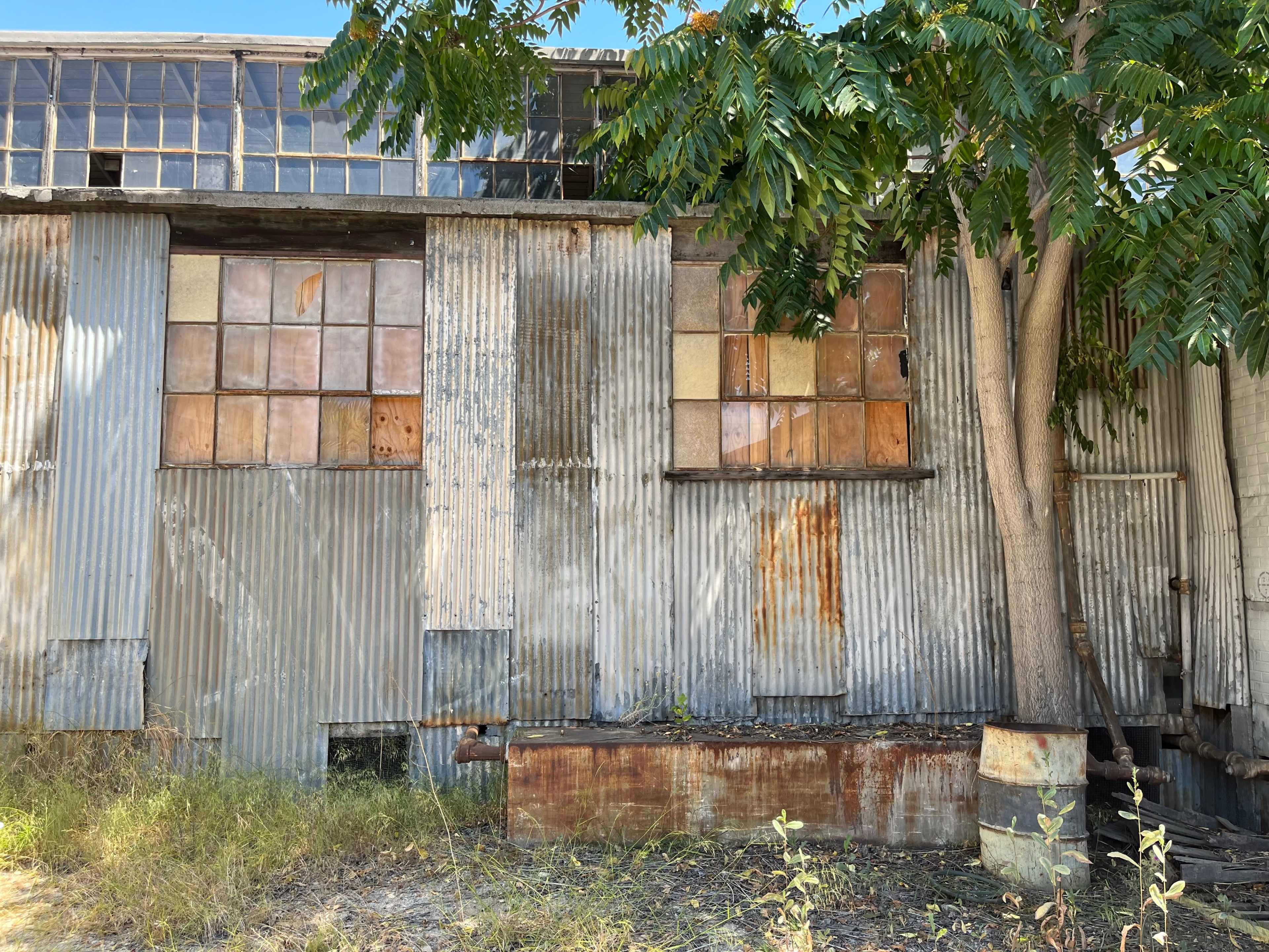 An old, weathered industrial building with corrugated metal siding and several boarded-up windows, partially obscured by a tree.