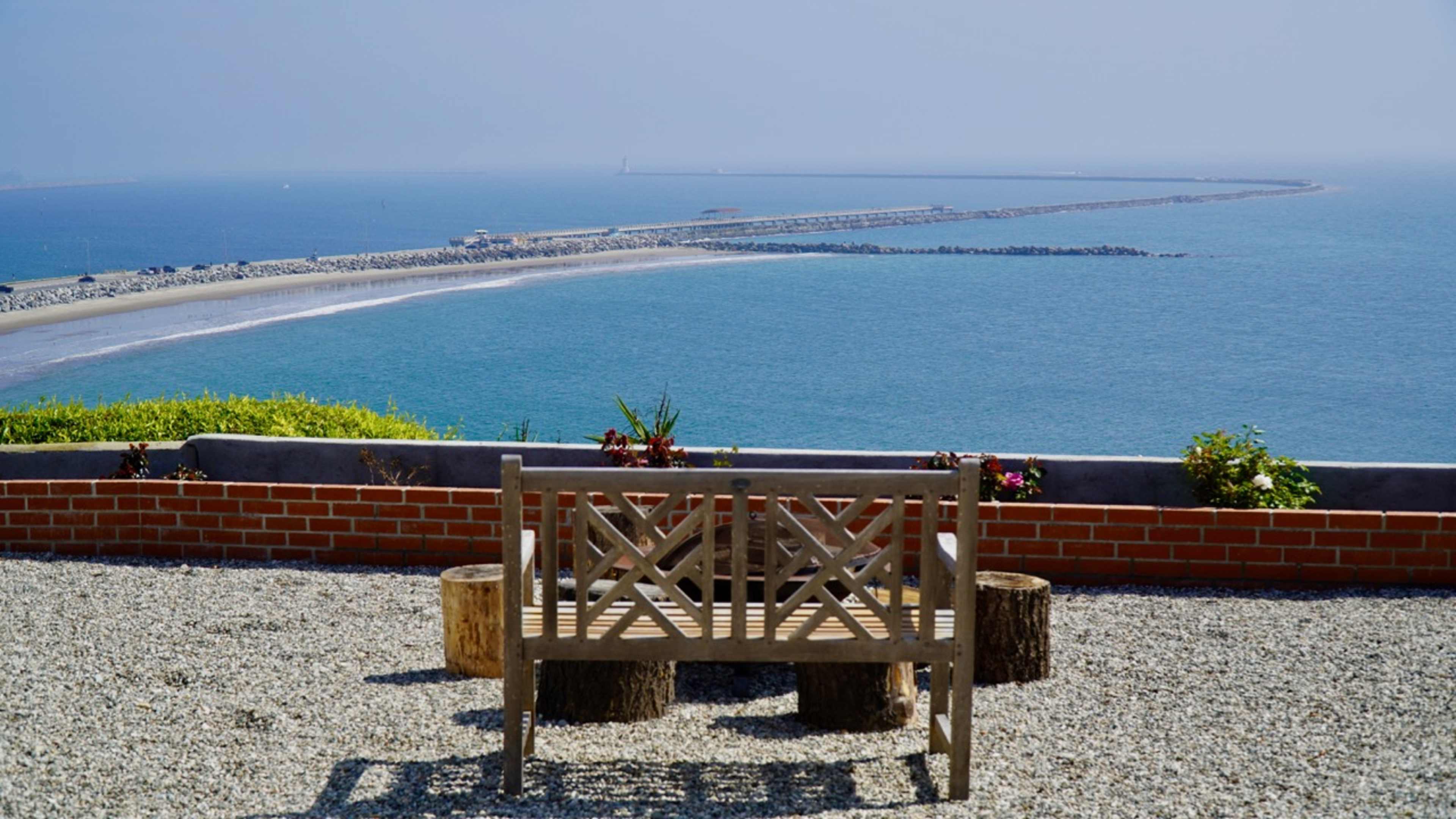 A wooden bench overlooks a rocky jetty extending into a calm blue sea under a clear sky.