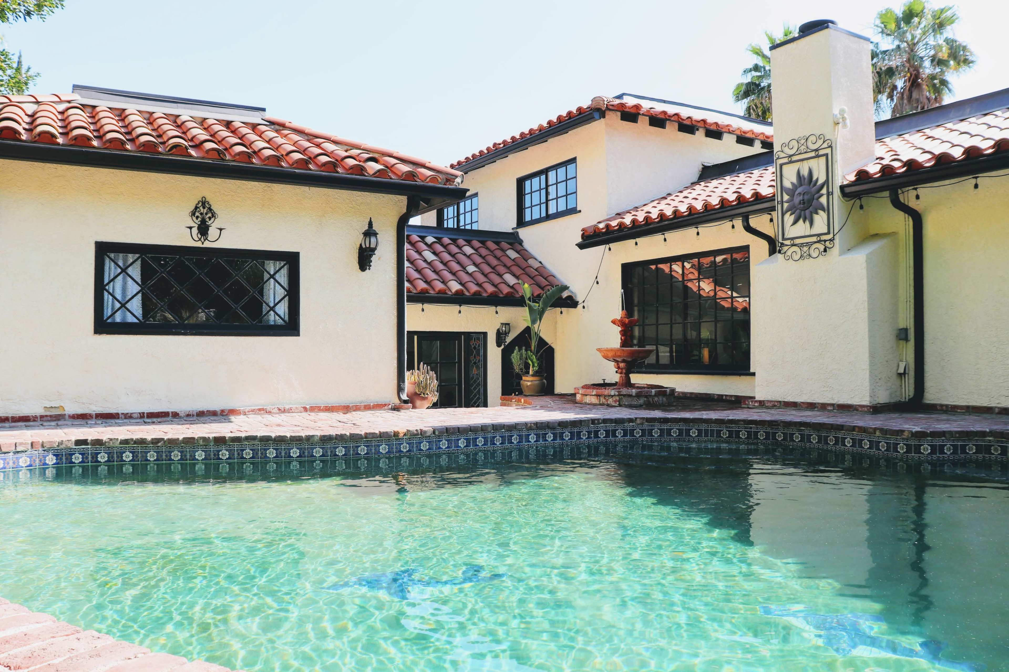 A clear swimming pool surrounded by a stucco home with red tile roofs and a fountain.
