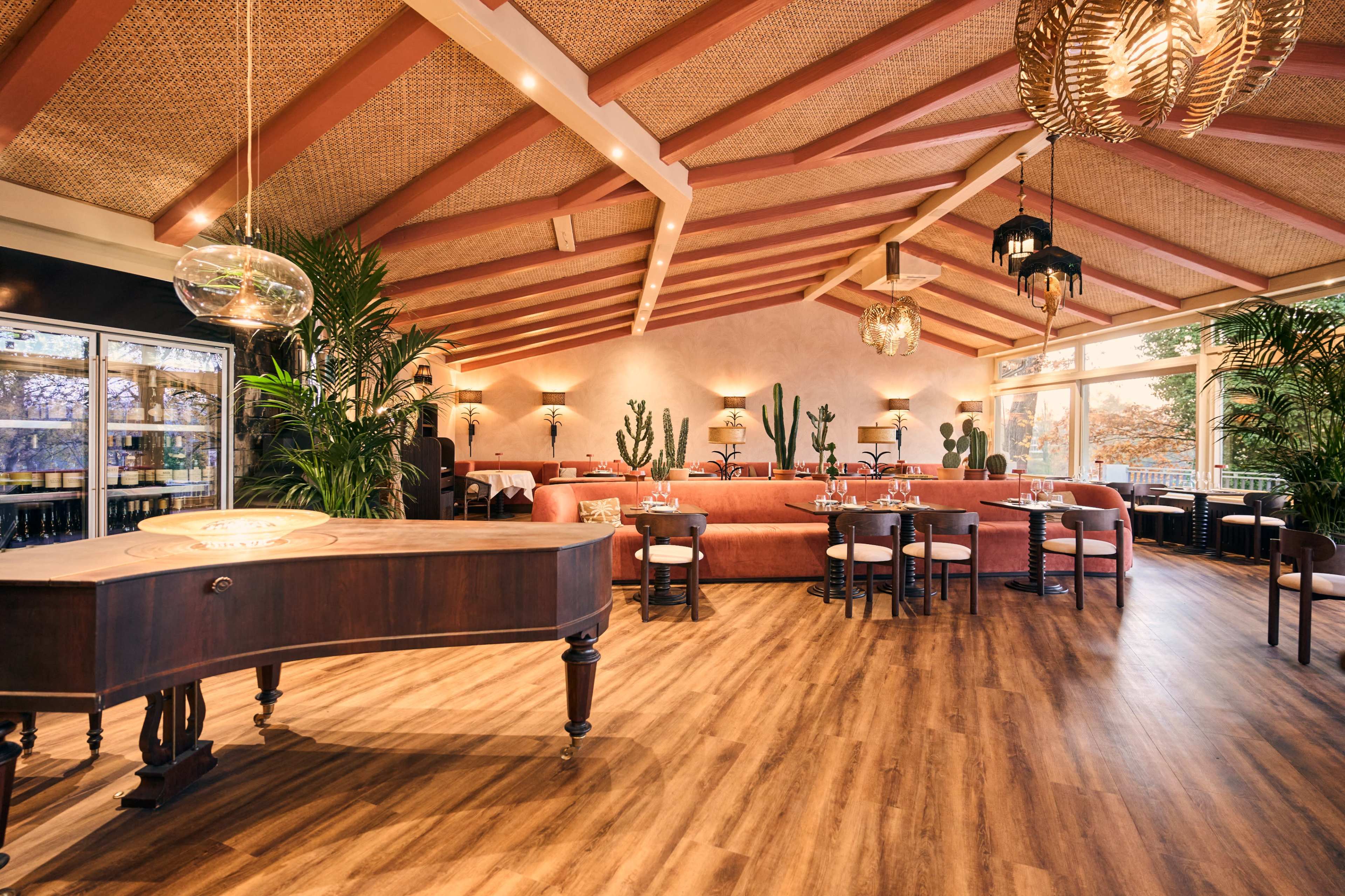 The image depicts a stylish restaurant interior featuring wooden floors, a high, beamed ceiling adorned with woven decor, and seating areas filled with plants.