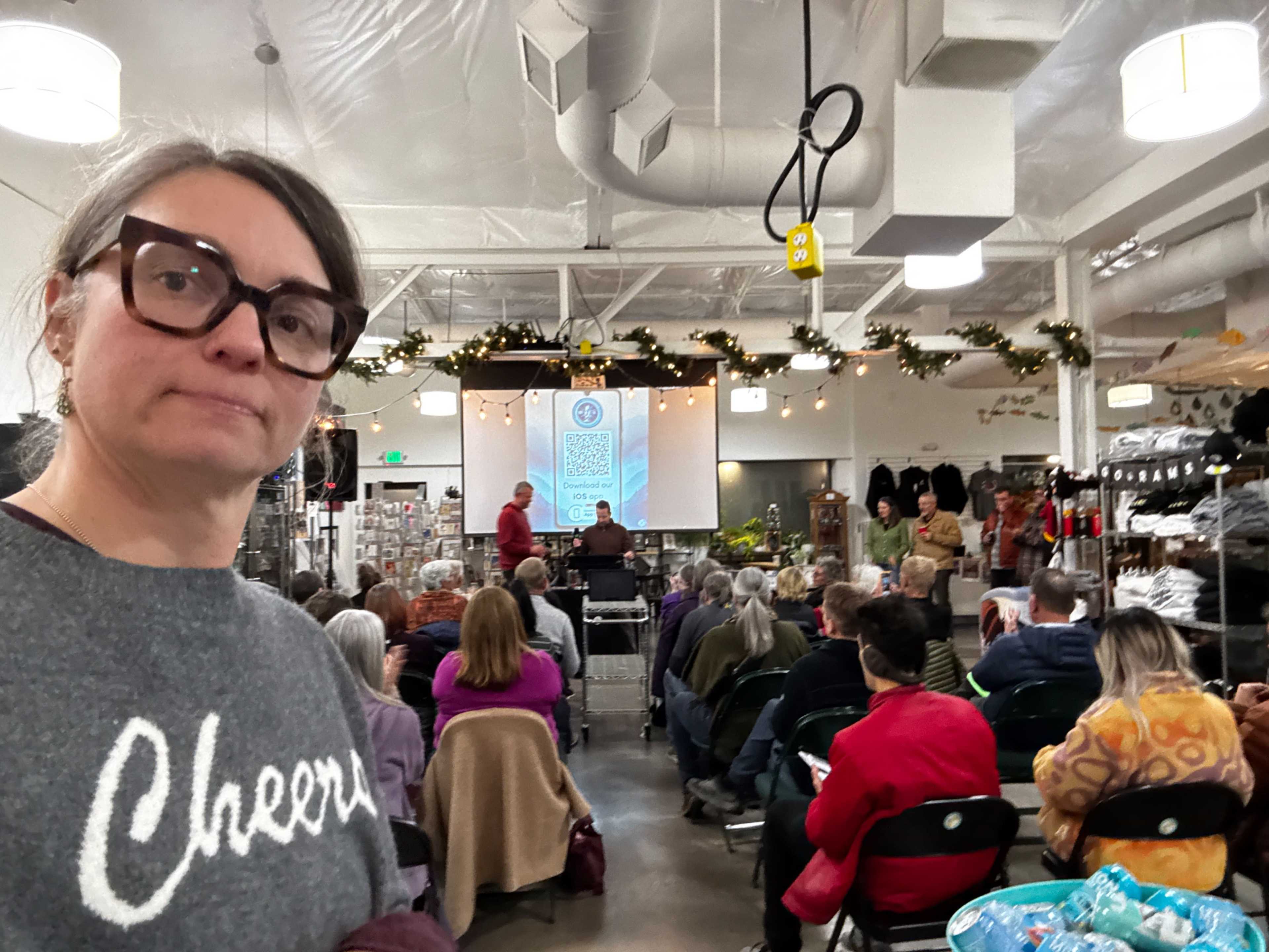 A woman takes a selfie in front of a crowd seated for a presentation in a warmly decorated indoor market.