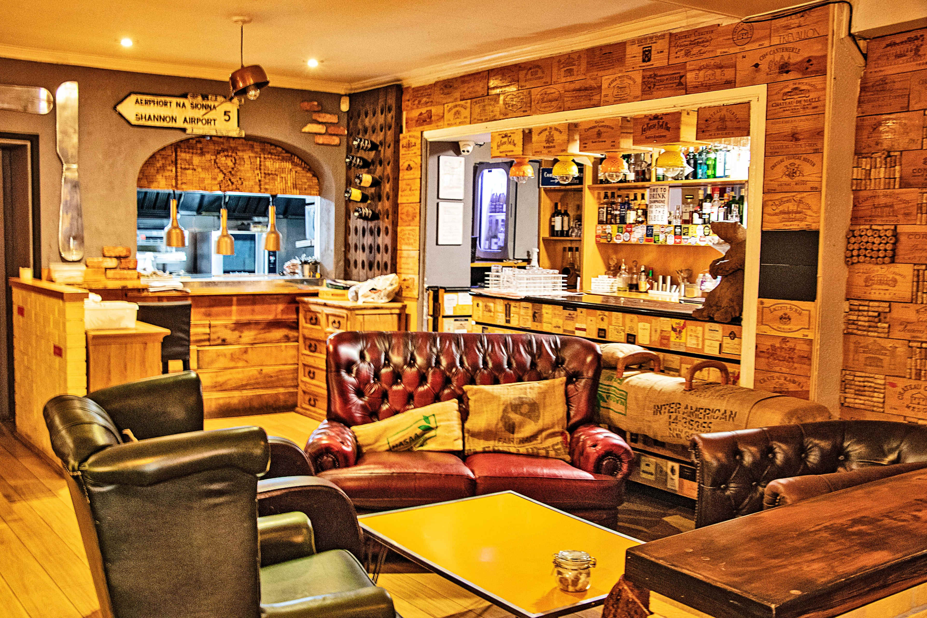 A cozy café interior featuring leather couches, wooden walls adorned with crates, and a counter displaying shelves of drinks and snacks.