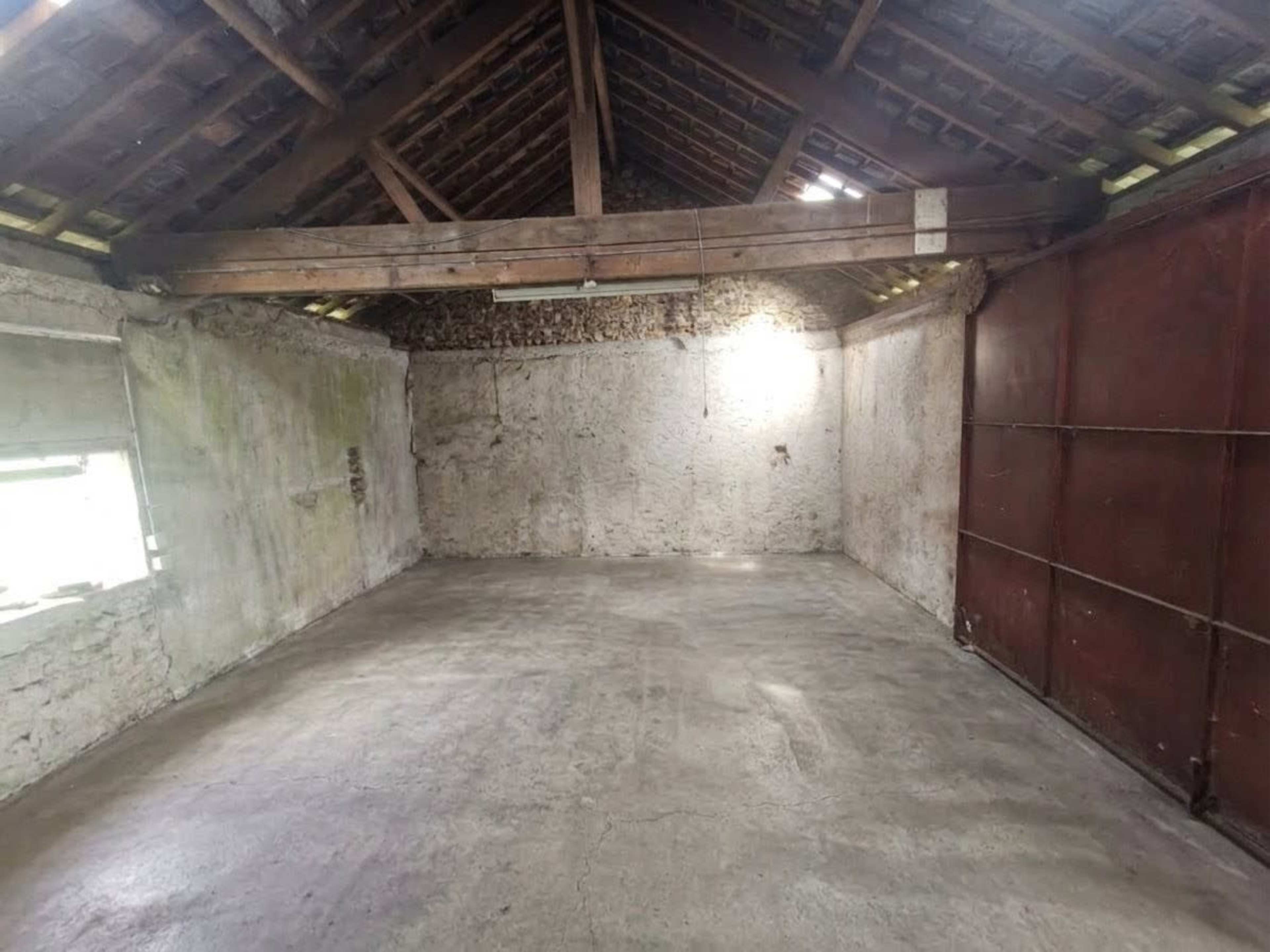 The image shows a large, empty room with a concrete floor, rough stone walls, and a high wooden ceiling.