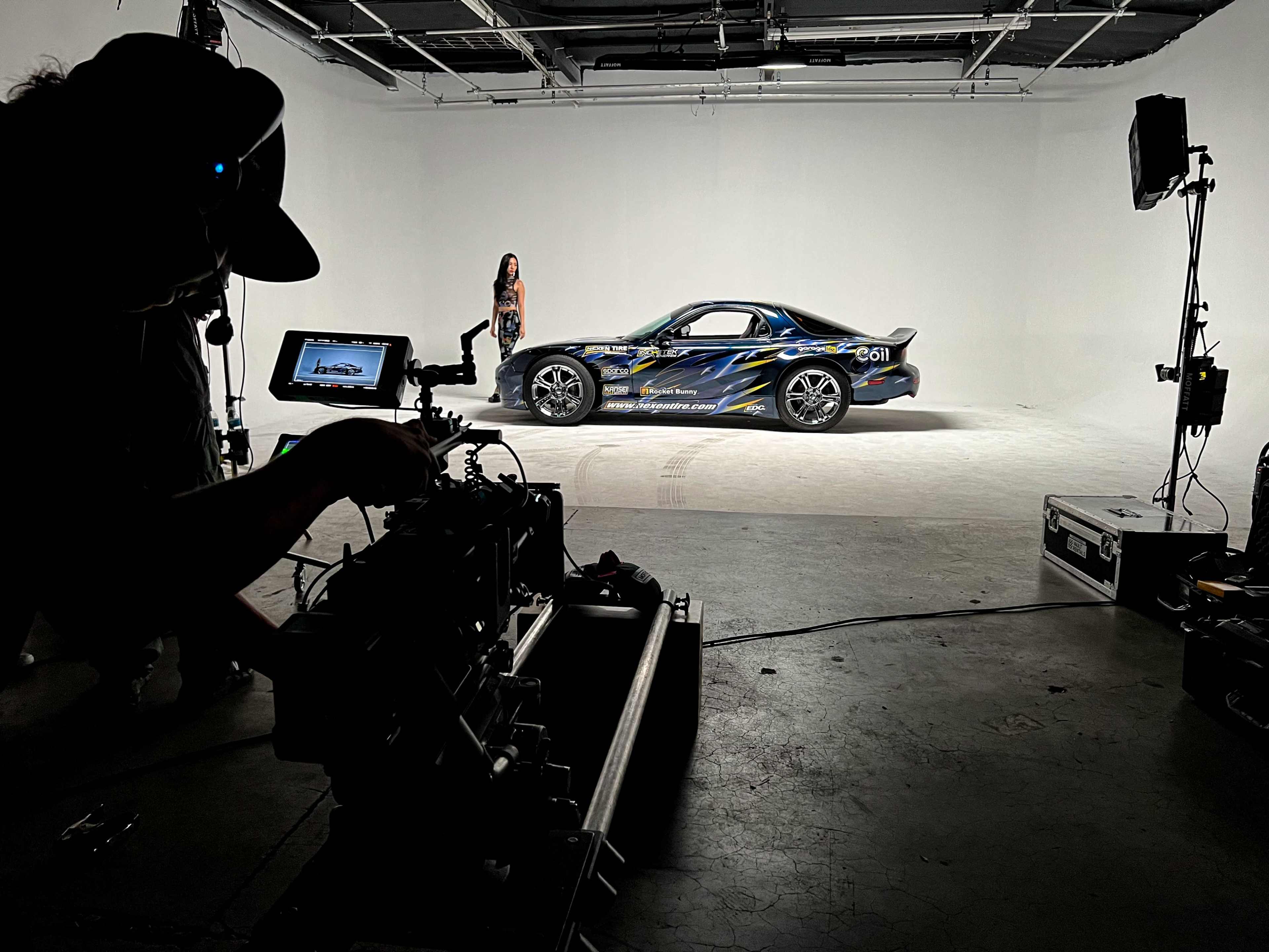 A film crew captures a model posing next to a decorated car in a studio setting.