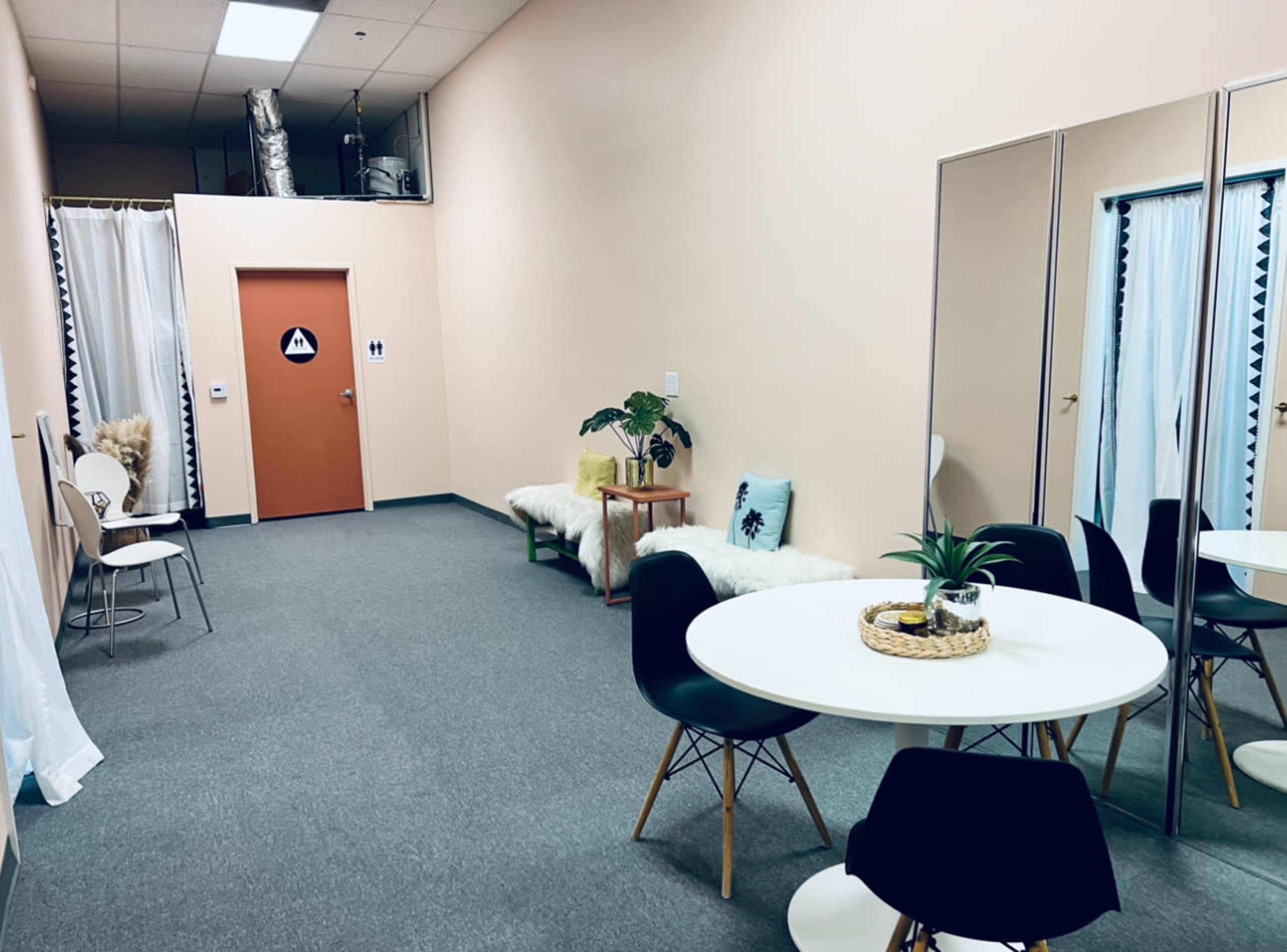 A small, minimalist waiting area with a round table, several chairs, a mirrored wall, and a decorated door leading to a restroom.