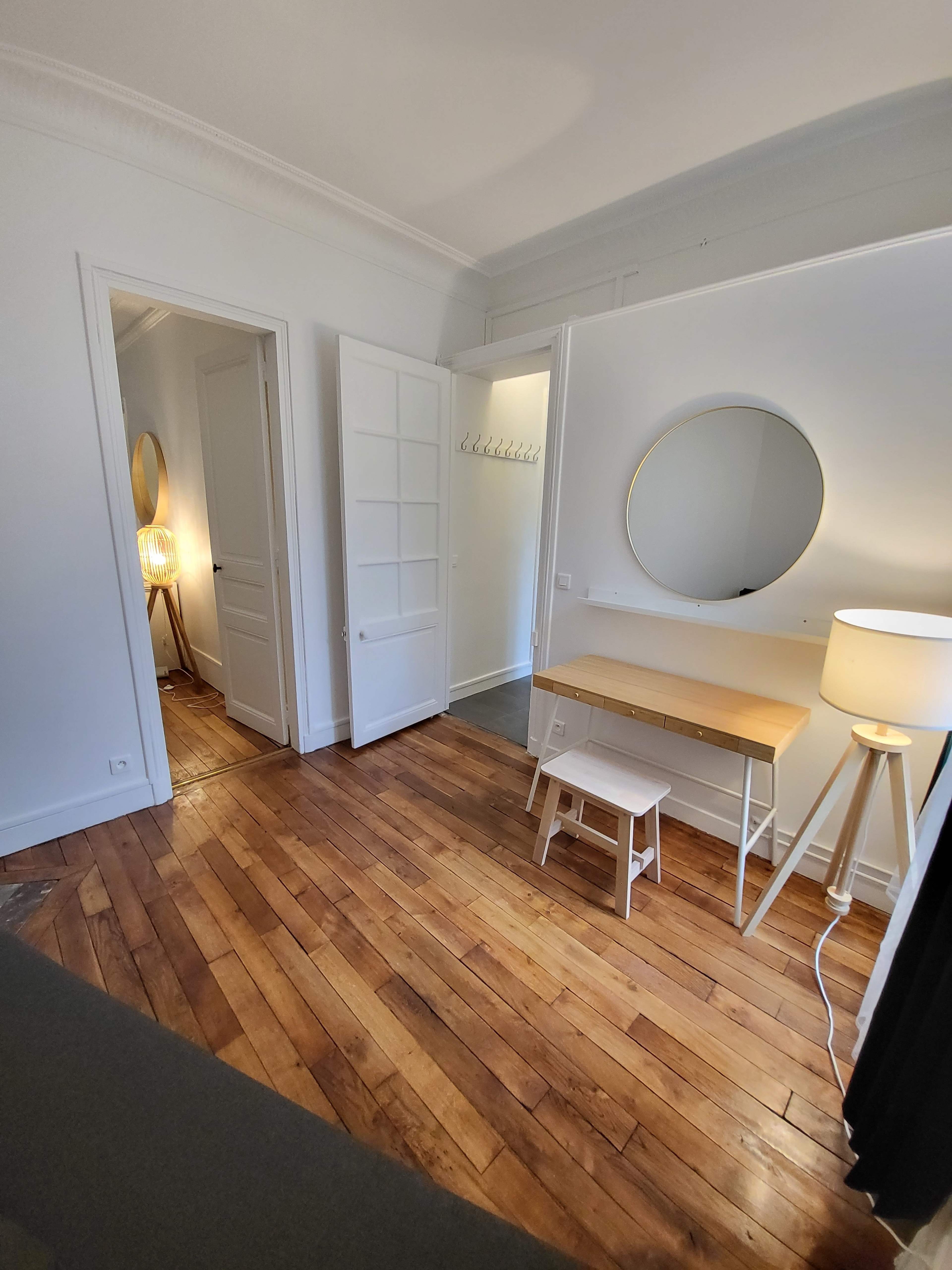 The image shows a well-lit room with wooden floors, featuring a circular mirror above a simple desk, a white stool, and a lamp beside an open doorway.