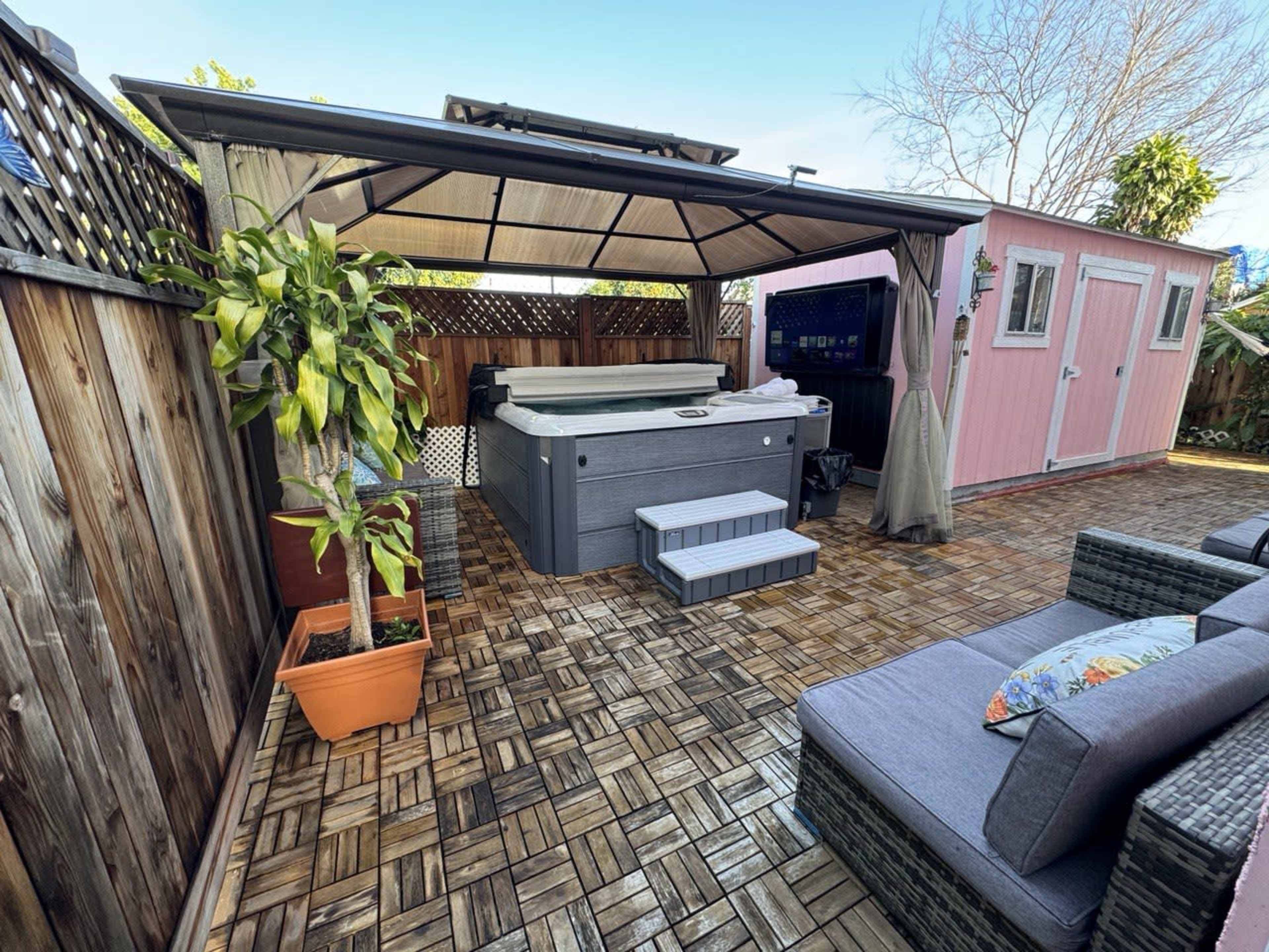 A backyard with a hot tub under a canopy, surrounded by wooden fencing, potted plants, and a pink storage shed.