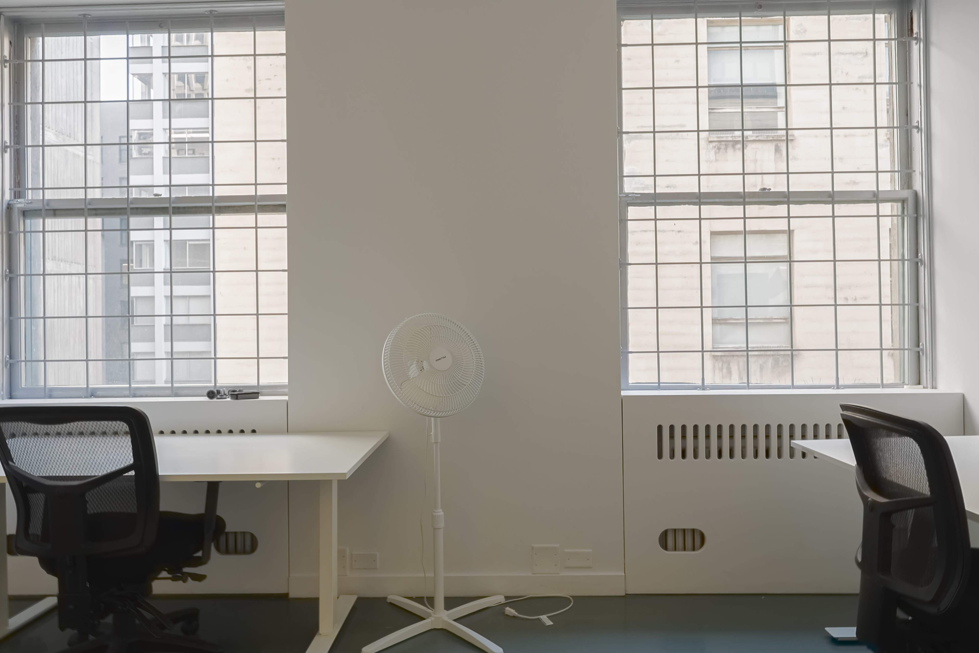 The image shows a minimalist office space featuring two empty desks facing windows with metal grills and a standing fan between them.