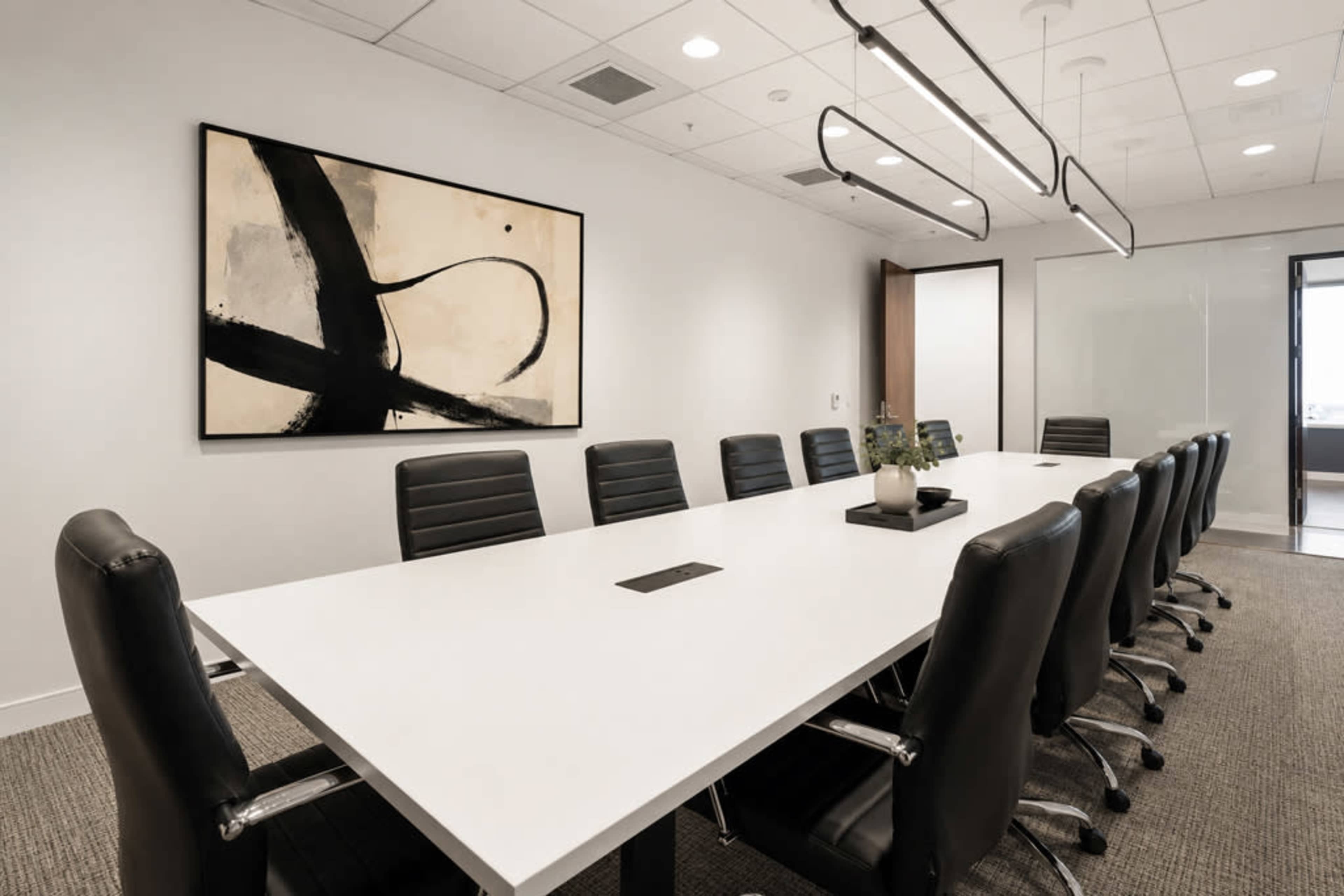 A modern conference room features a long white table surrounded by black leather chairs, with a large abstract painting on the wall and a small plant centerpiece on the table.